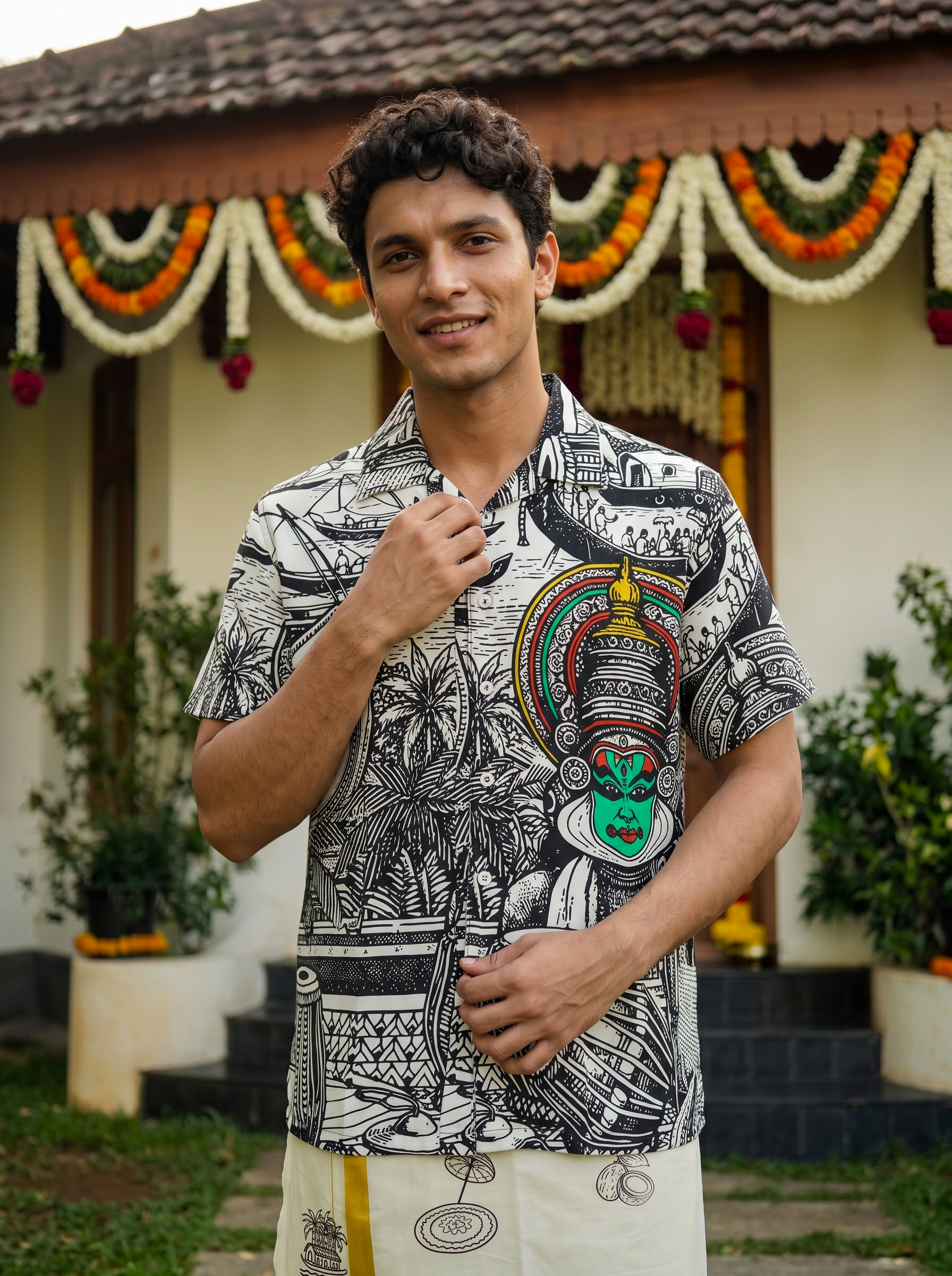 A man wearing a black and white graphic print short-sleeve button-up shirt with a colorful traditional mask design on the chest, standing outdoors in front of a decorated building.