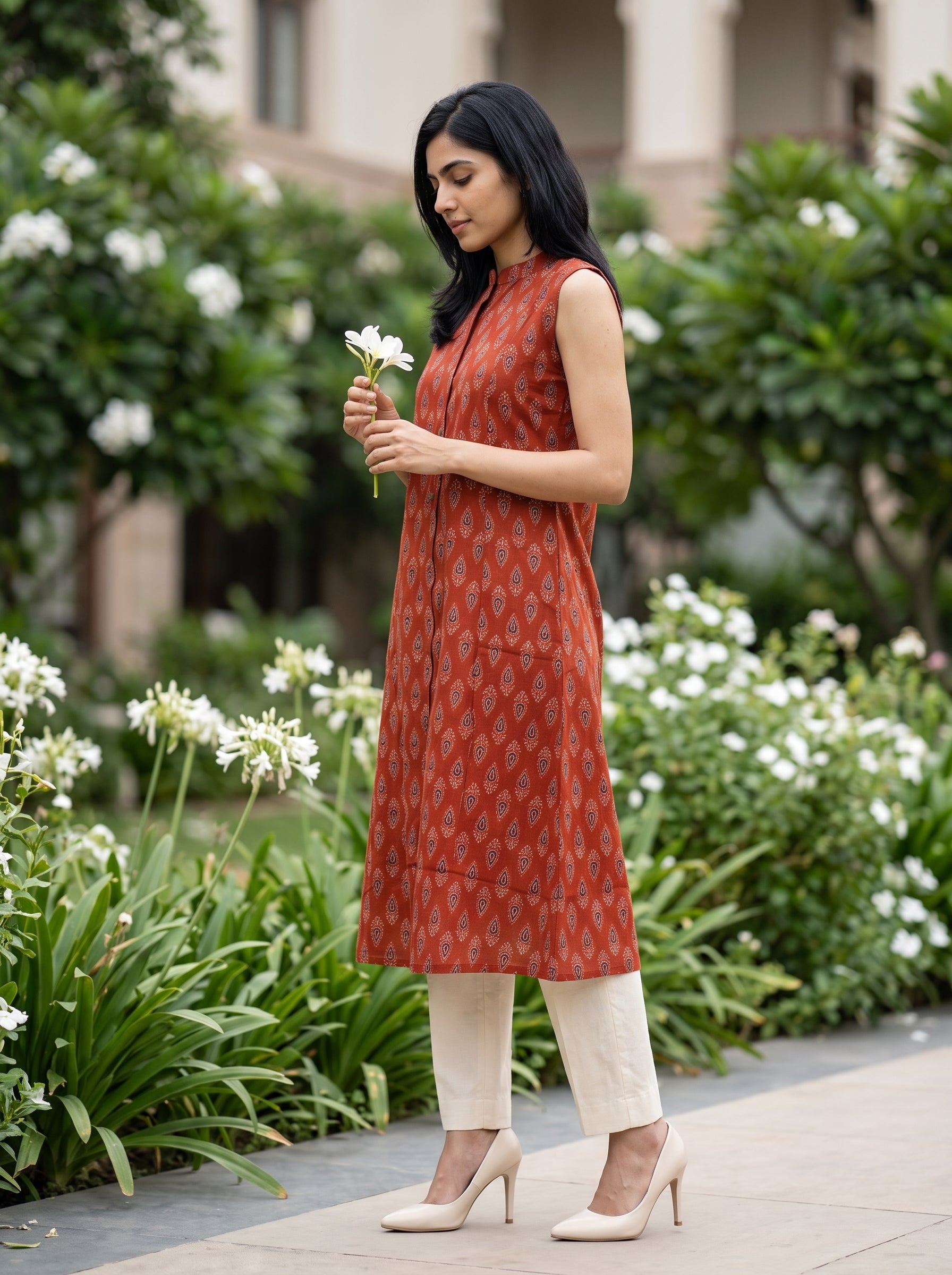 A woman stands outdoors in a garden setting wearing a sleeveless rust-orange printed kurta with cream pants and beige heels, holding a small white flower.