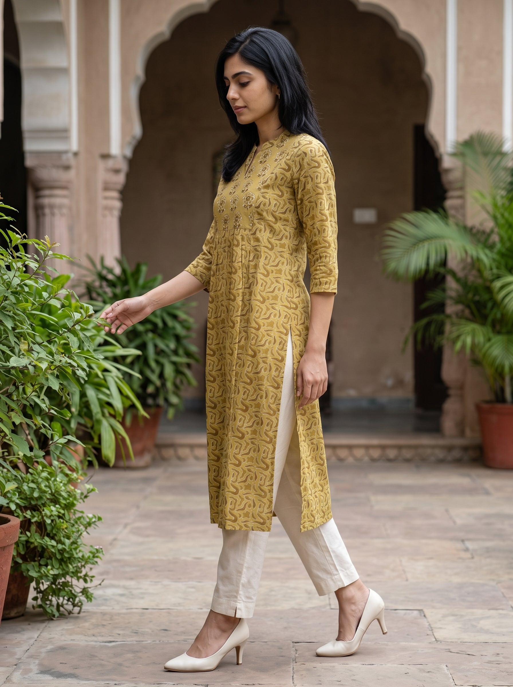 A woman wearing a mustard yellow kurta with a subtle brown pattern, paired with off-white trousers and white heels, standing in a courtyard with arches and potted plants.