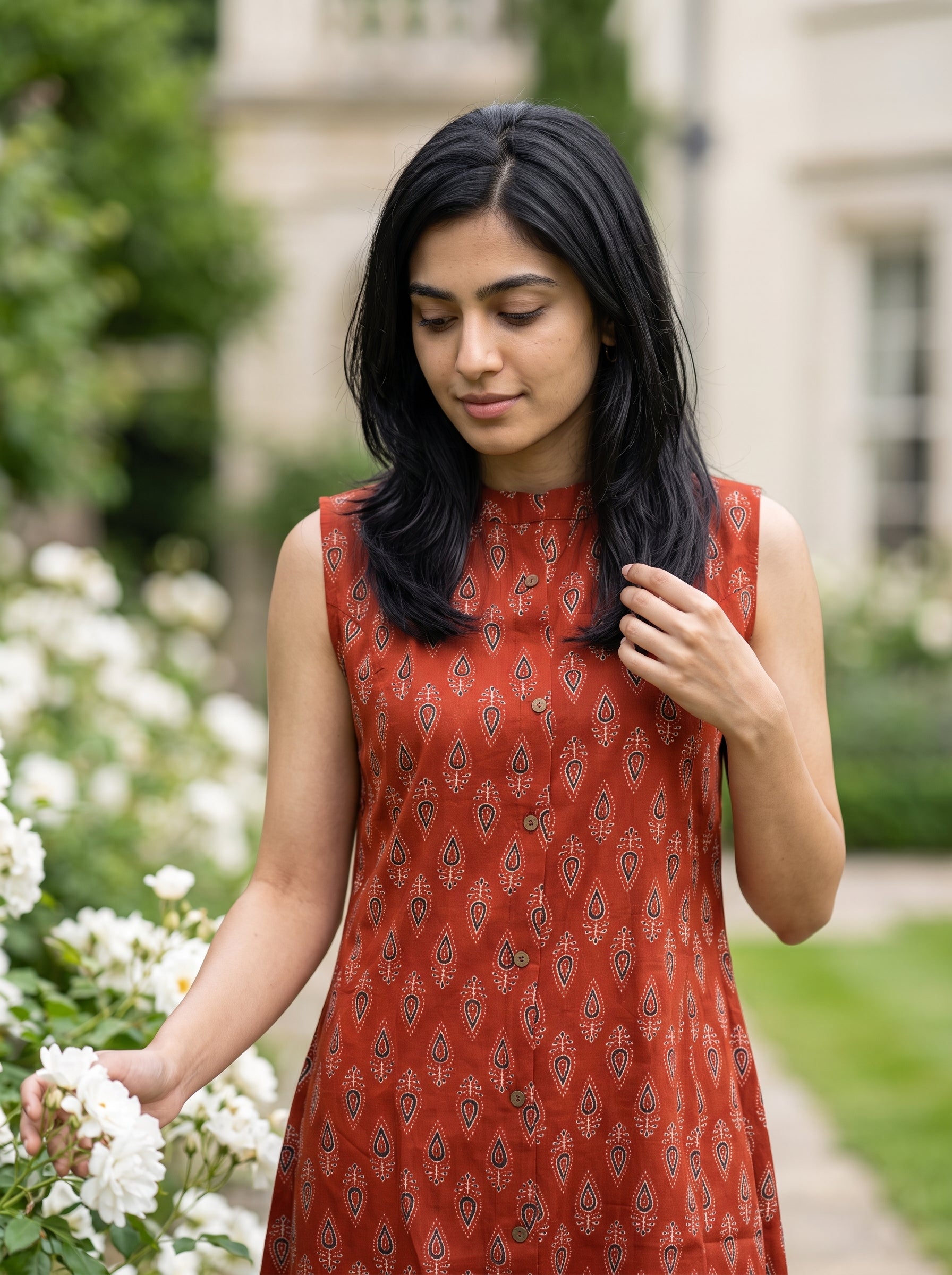 A woman wearing a sleeveless rust-orange dress with a repeating dark geometric pattern, standing in a garden next to white flowers.