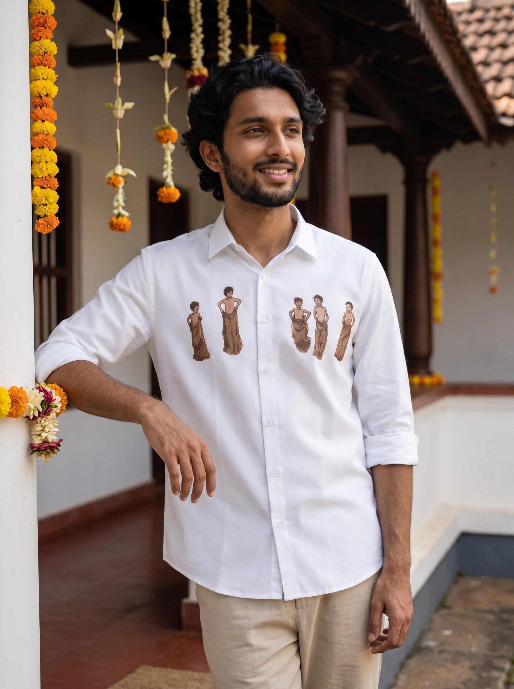 A man wearing a white long-sleeve button-up shirt with embroidered figures on the chest, paired with beige trousers, standing in a traditional courtyard decorated with marigold garlands.