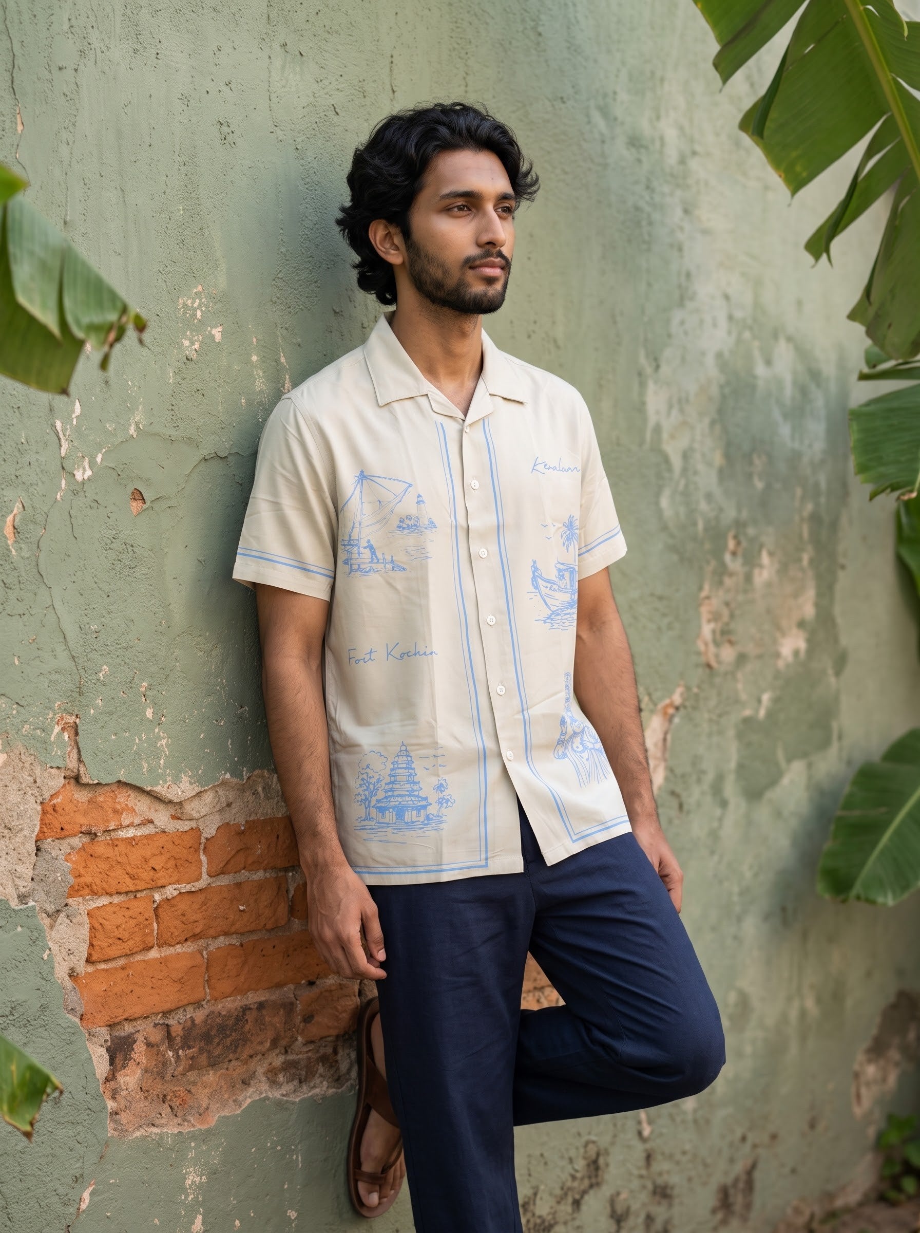 A man wearing a cream-colored short-sleeve button-up shirt with light blue illustrations and navy blue trousers, leaning against a weathered green wall with exposed brick and tropical leaves.