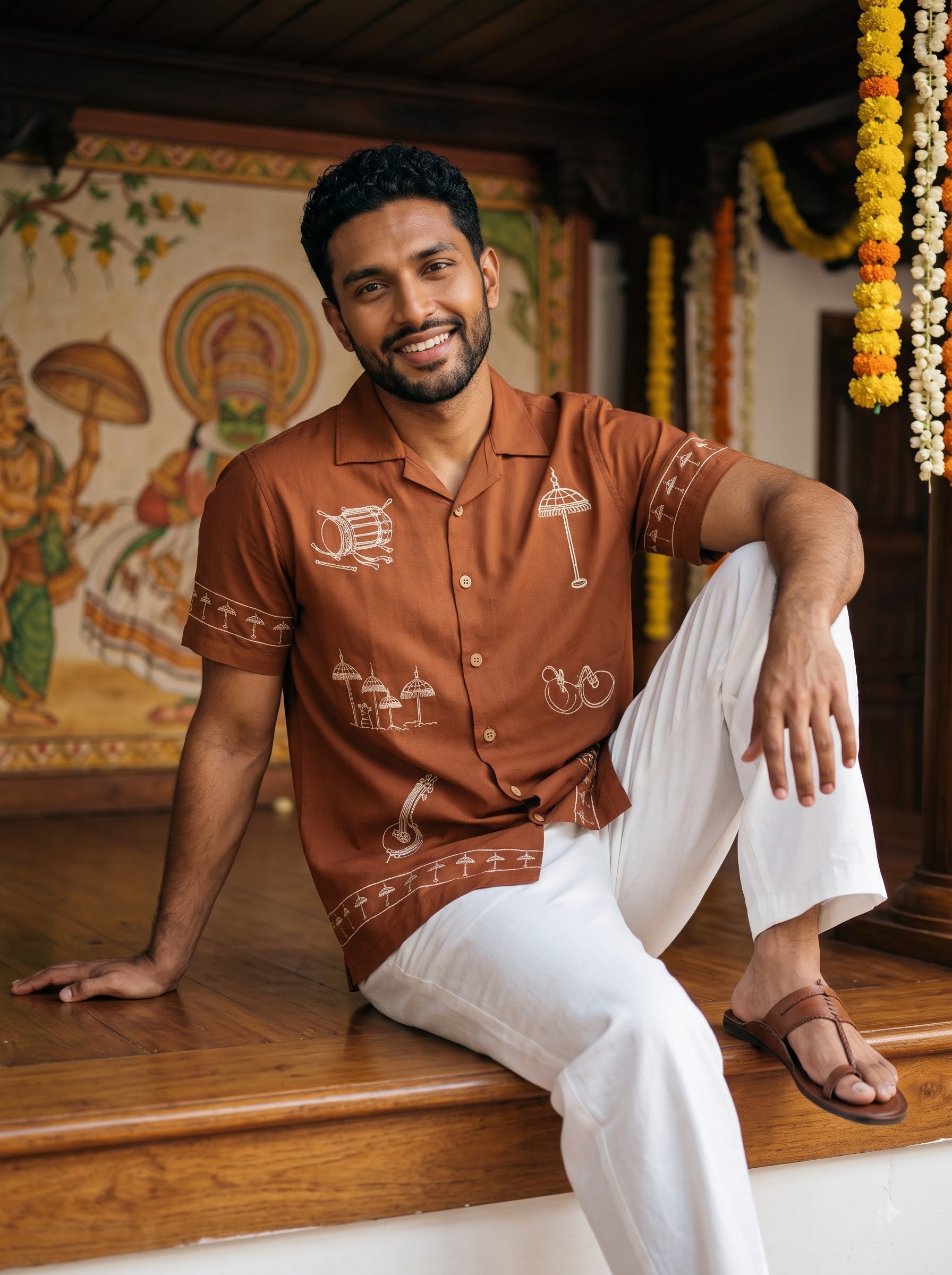 A man wearing a rust-brown short-sleeve button-up shirt with white printed motifs and white trousers, sitting on a wooden ledge in a decorated indoor setting.