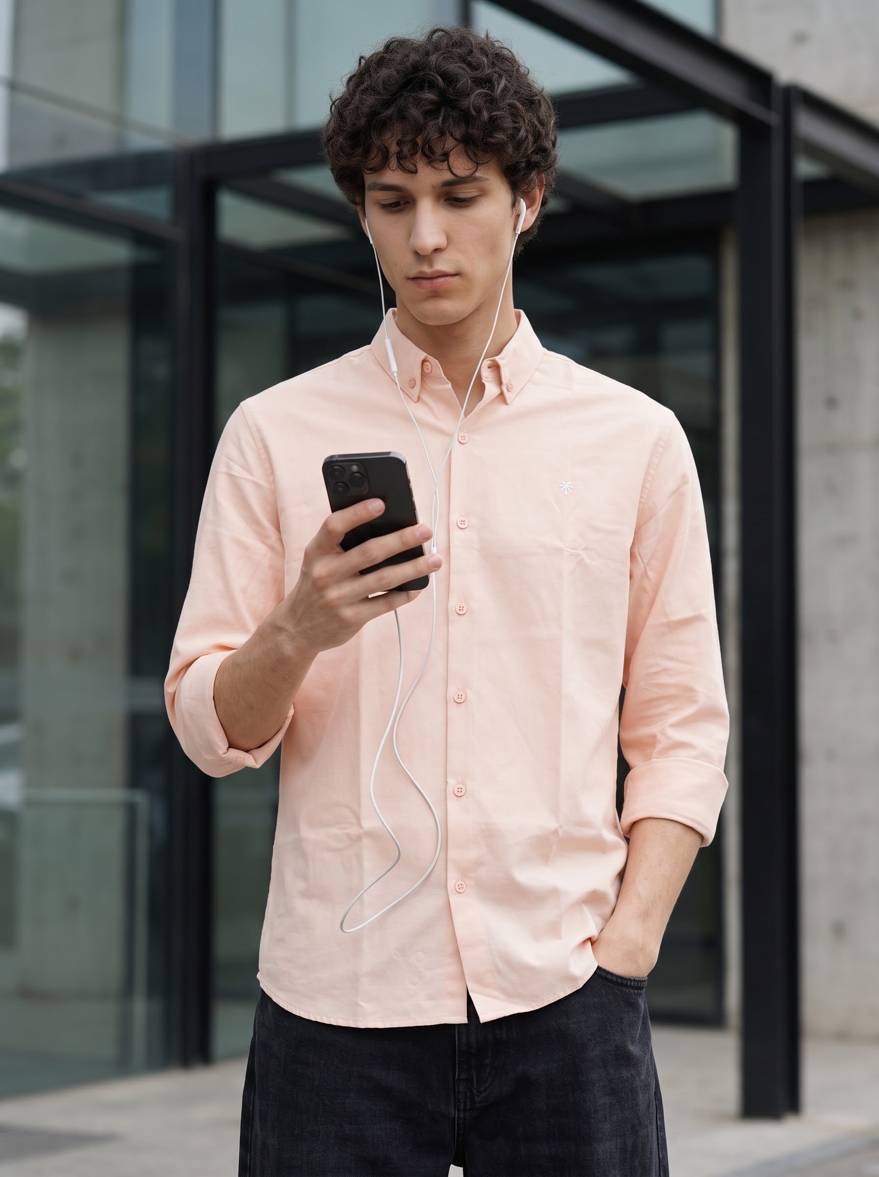 A man wearing a light peach-colored long-sleeve button-down shirt and dark jeans, looking down at a black smartphone he is holding. White wired earphones are plugged into his ear, with a modern glass and concrete building in the background.