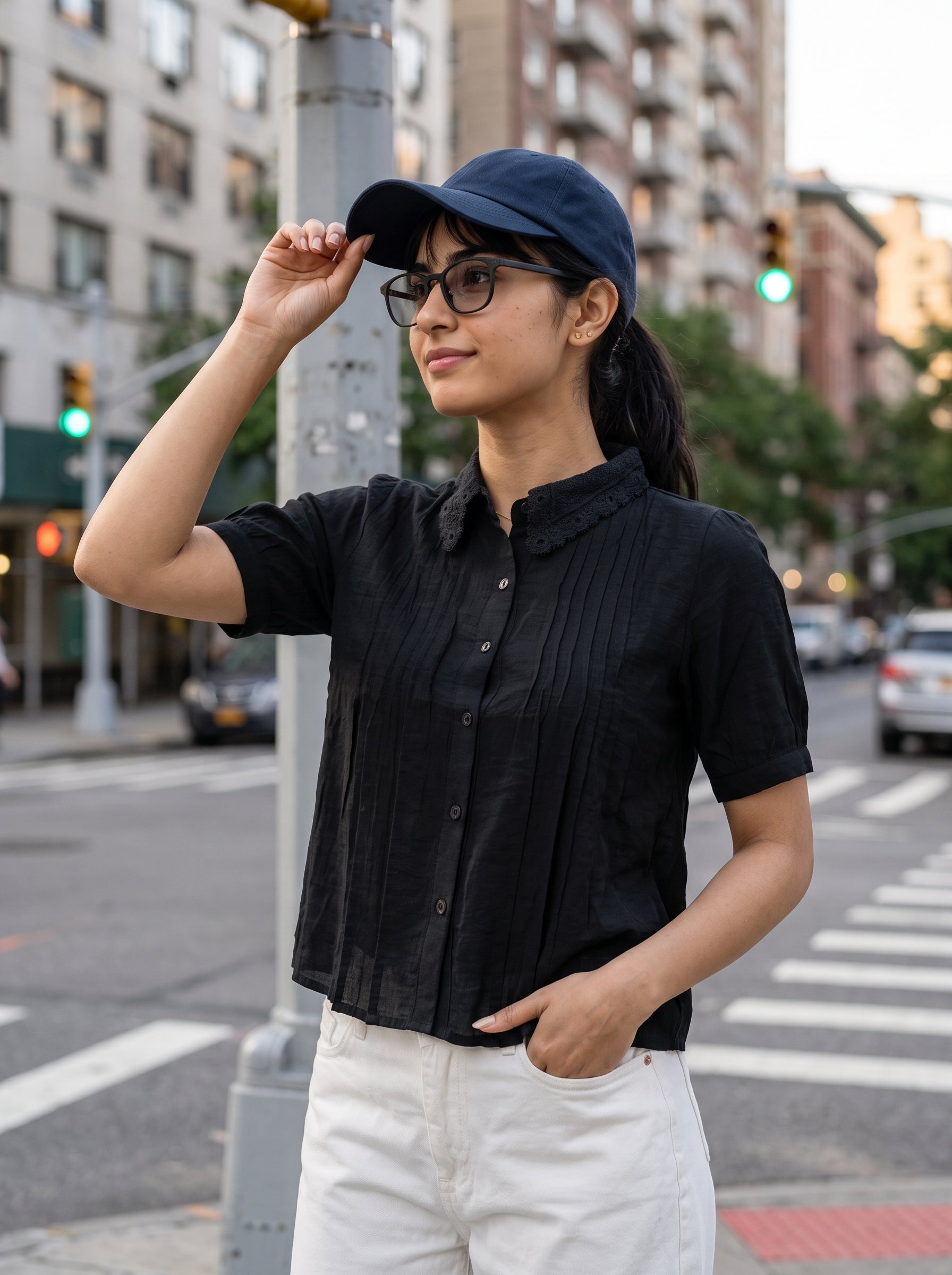 A woman standing on a city street crosswalk wearing a black short-sleeve button-up shirt, white pants, a navy blue baseball cap, and glasses.
