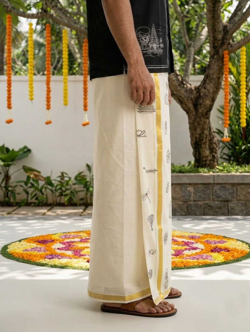 A person wearing a traditional Indian dhoti or lungman in off-white with a gold border and printed motifs, paired with a black shirt and sandals, standing outdoors in a garden courtyard with flower garlands and a floral rug on the ground.
