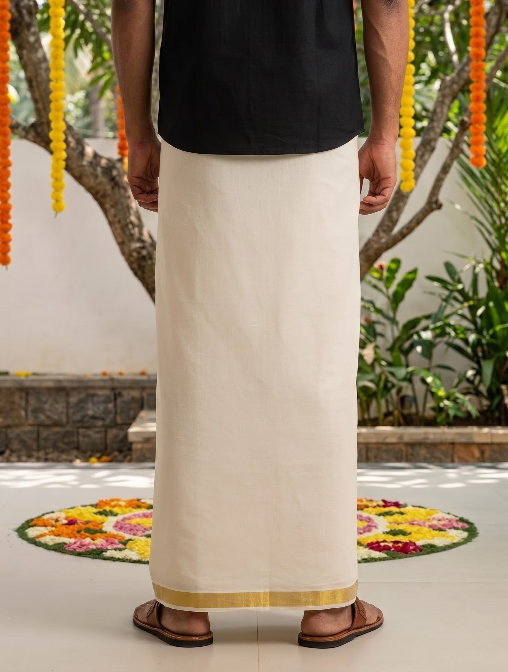 A person shown from the back wearing a long off-white traditional wrap skirt with a thin gold border, paired with a black top and brown sandals, standing outdoors in front of a decorated wall with hanging flower garlands.