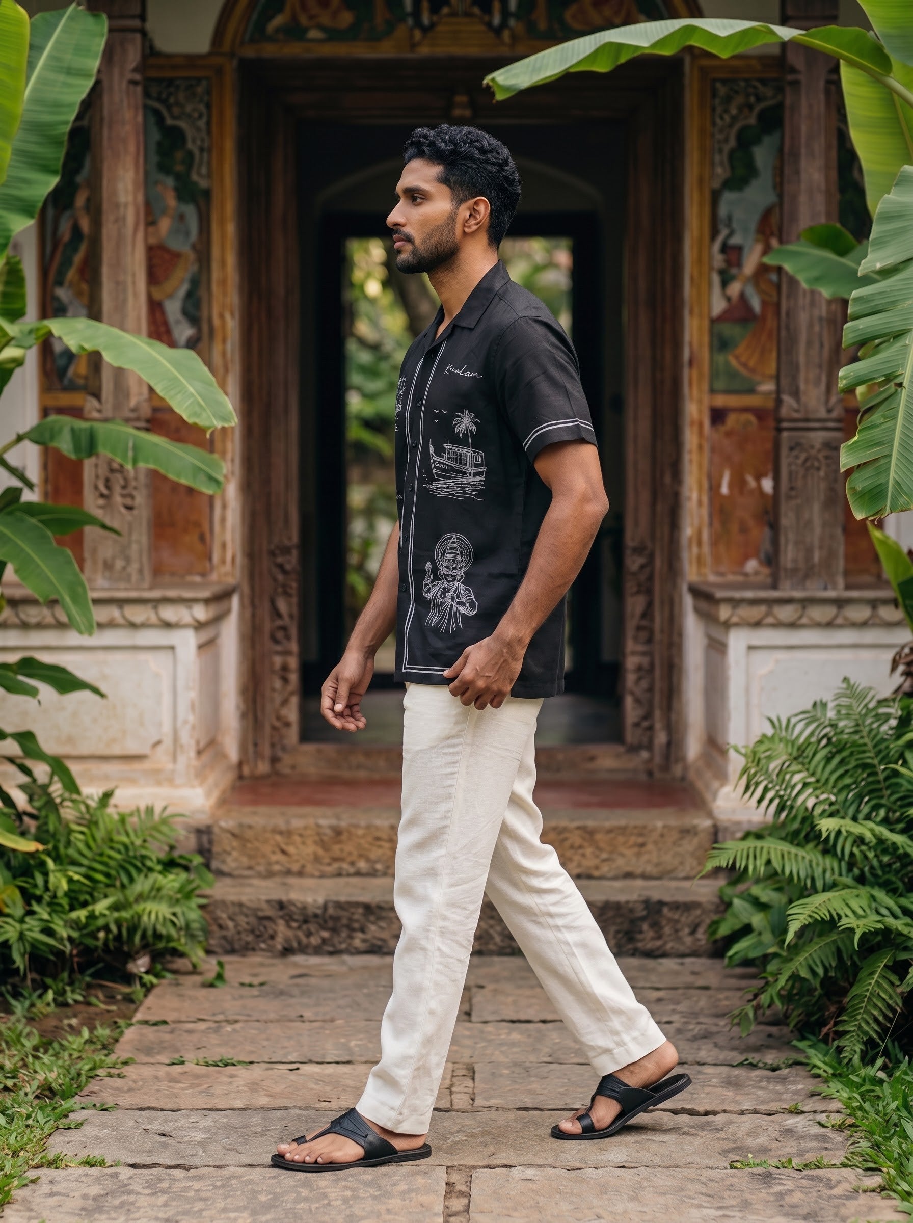 A man wearing a black short-sleeve button-up shirt with white graphics, off-white trousers, and black sandals walks along a stone path in front of a traditional carved wooden doorway surrounded by lush green plants.