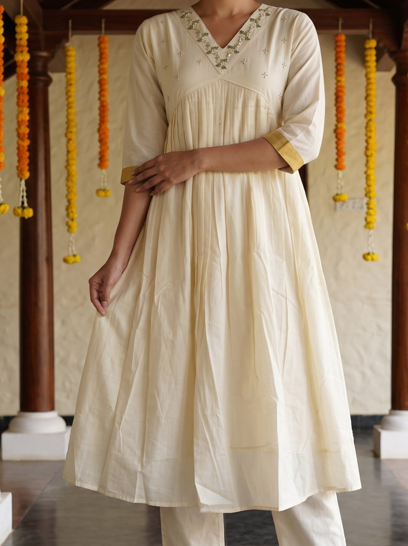 A woman wearing a cream-colored cotton kurta with three-quarter sleeves, a V-neckline with delicate embroidery, and a flared skirt, paired with matching cream trousers. She is standing in a traditional corridor decorated with yellow and orange marigold garlands.