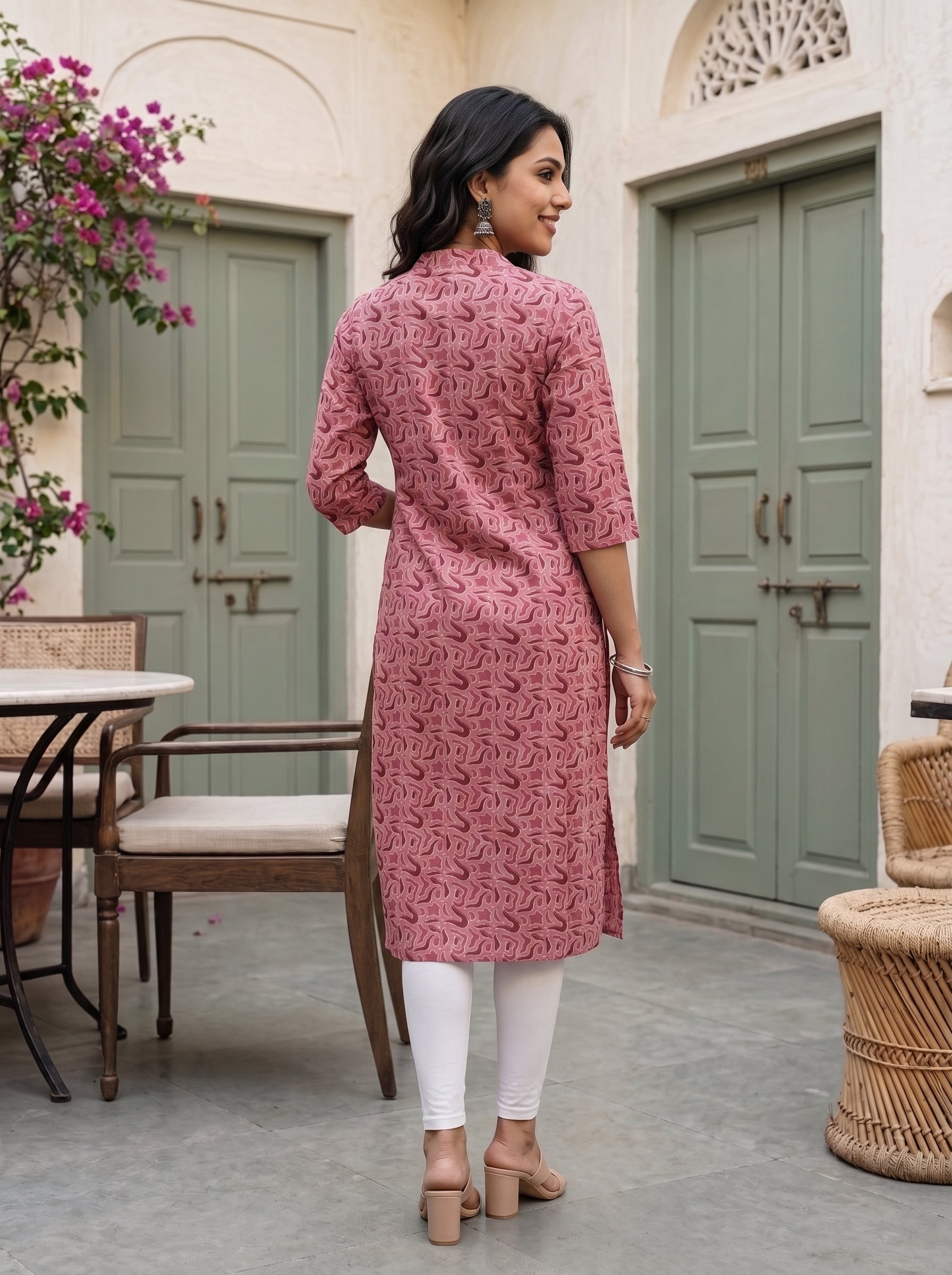 A woman wearing a pink printed kurta with three-quarter sleeves and white leggings, standing in a courtyard with green doors and wicker furniture.