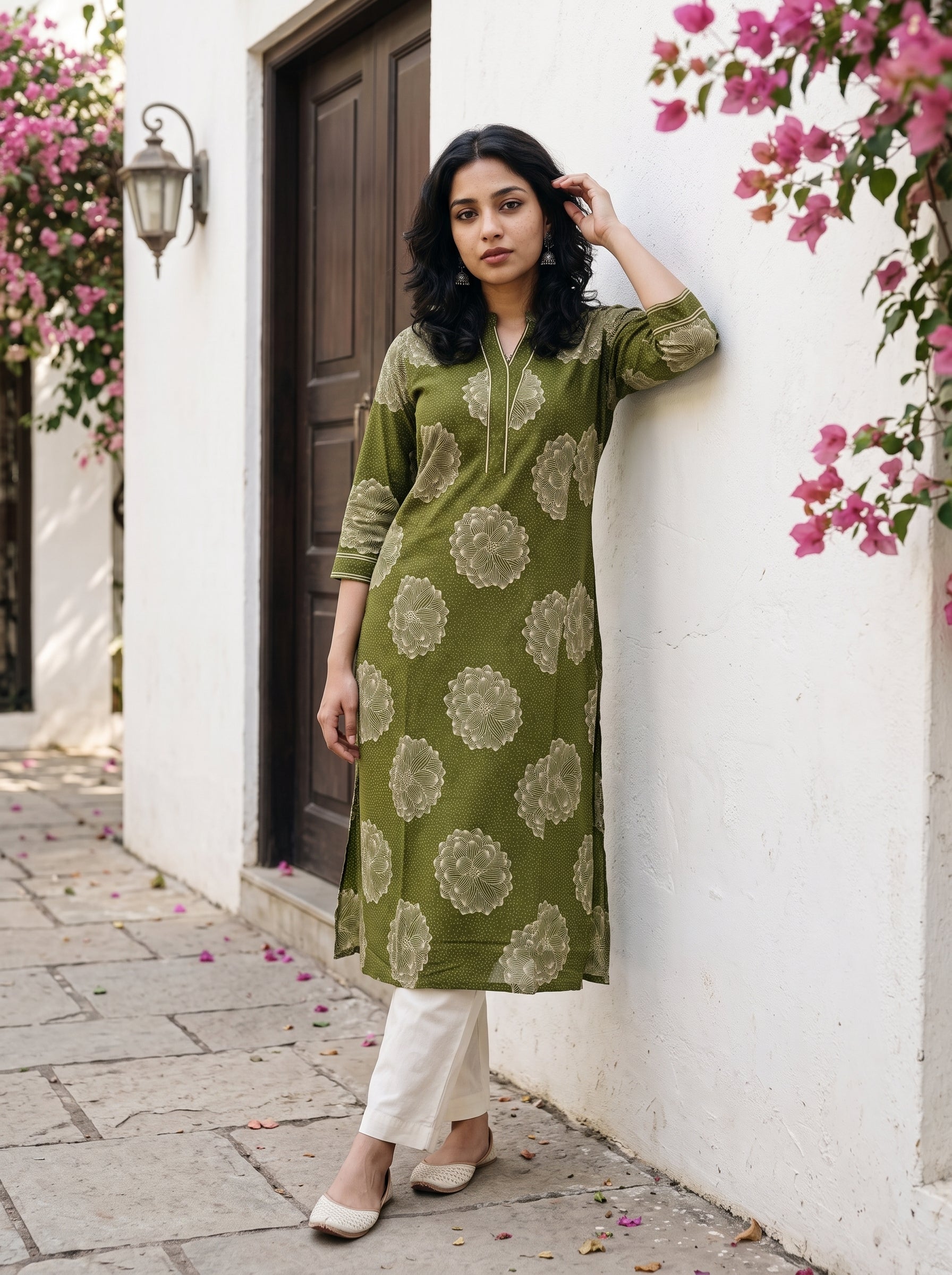 A woman stands outdoors against a white wall with pink flowers, wearing an olive green kurta with beige floral motifs and white pants.