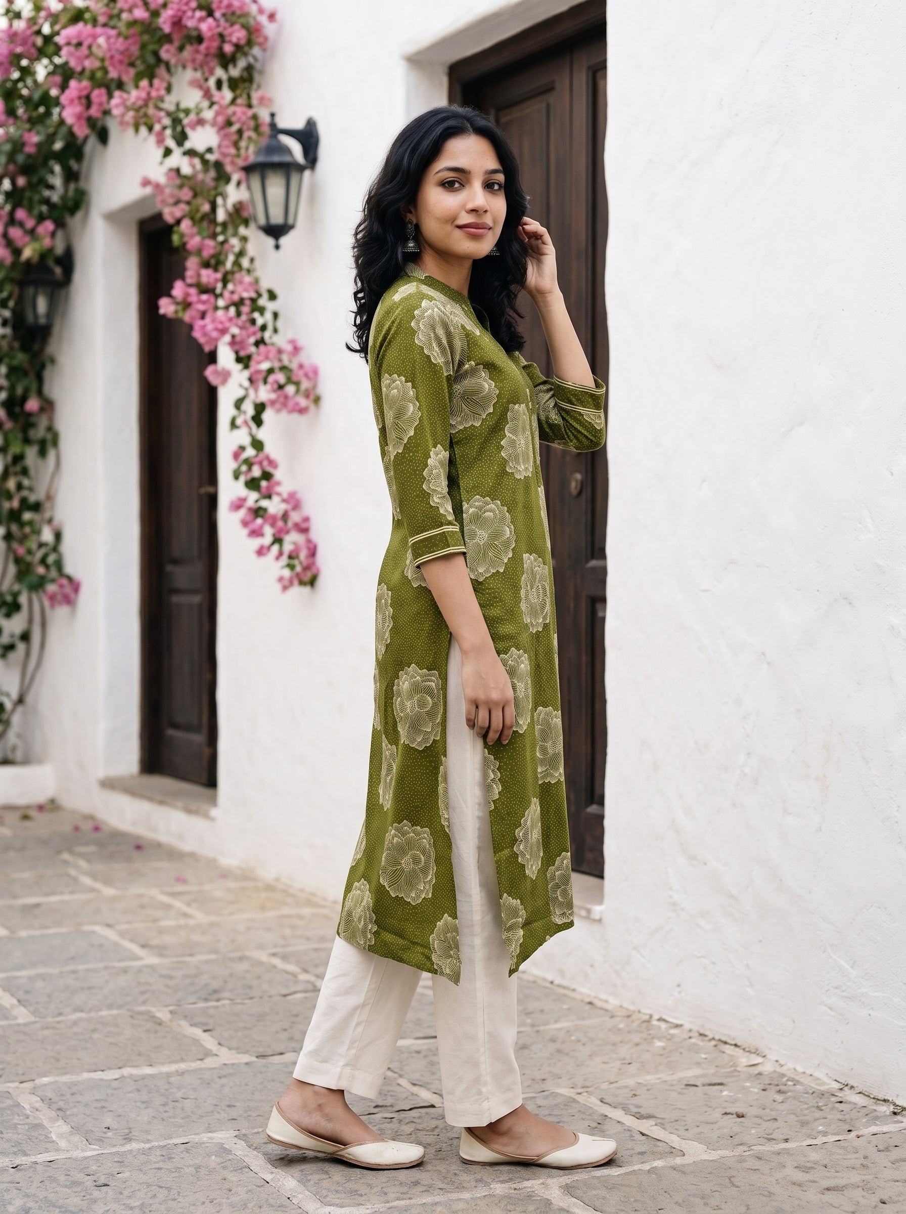 A woman stands outdoors in front of a white wall with dark wooden doors and pink flowers, wearing a green printed kurta with white pants and beige flat shoes.