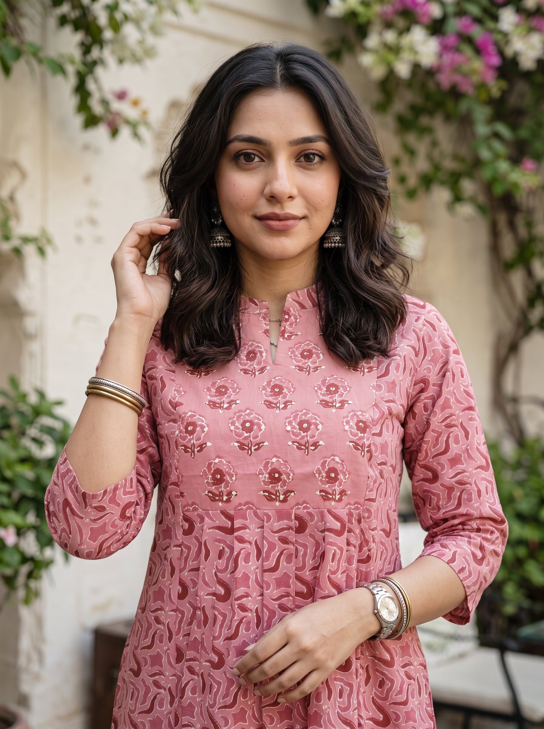 A woman wearing a pink embroidered kurta with floral patterns and 3/4 sleeves, standing outdoors in front of a light-colored wall with green foliage and flowers.