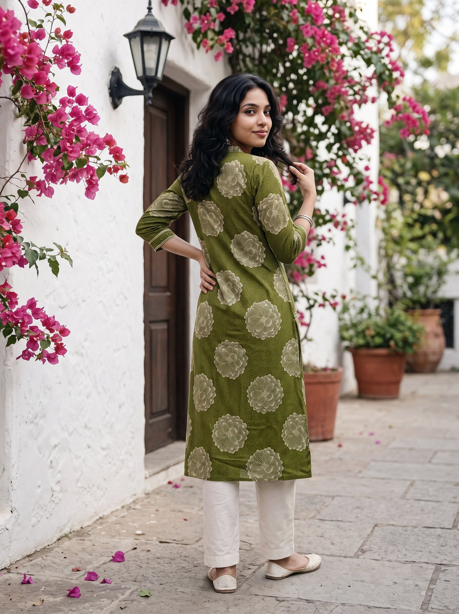 A woman wearing a green kurta with beige floral prints and white pants, standing on a stone pathway in front of a white wall with pink bougainvillea flowers.