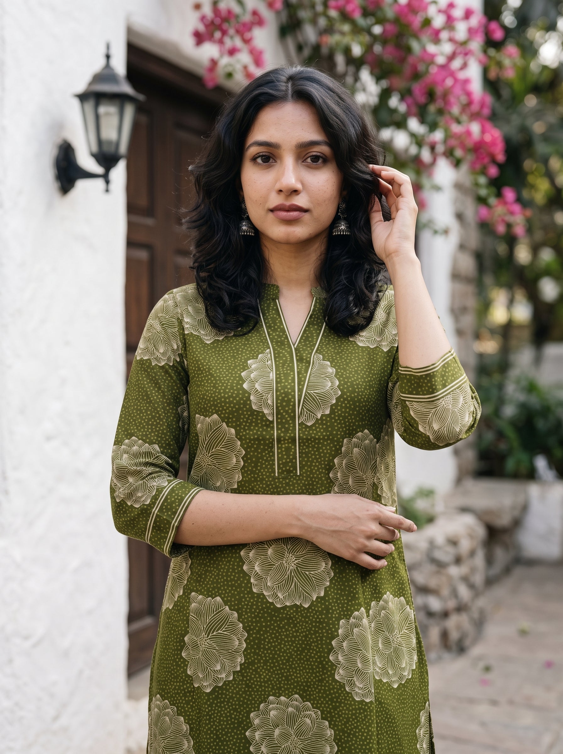 A woman wearing an olive green kurta with beige floral patterns, standing outdoors in front of a white wall and wooden door.