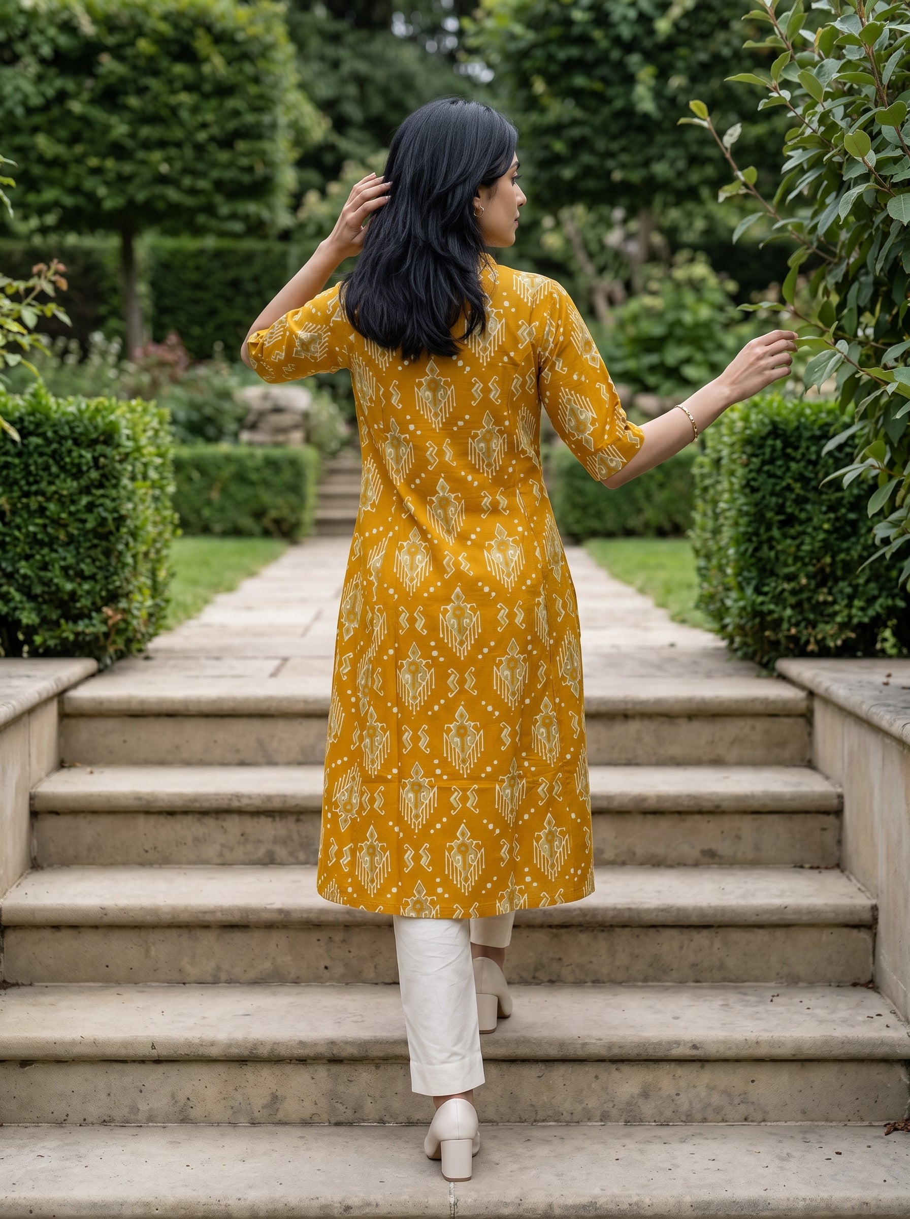 A woman viewed from behind wearing a mustard yellow and white patterned kurta with white pants, walking up stone steps in a lush green garden.