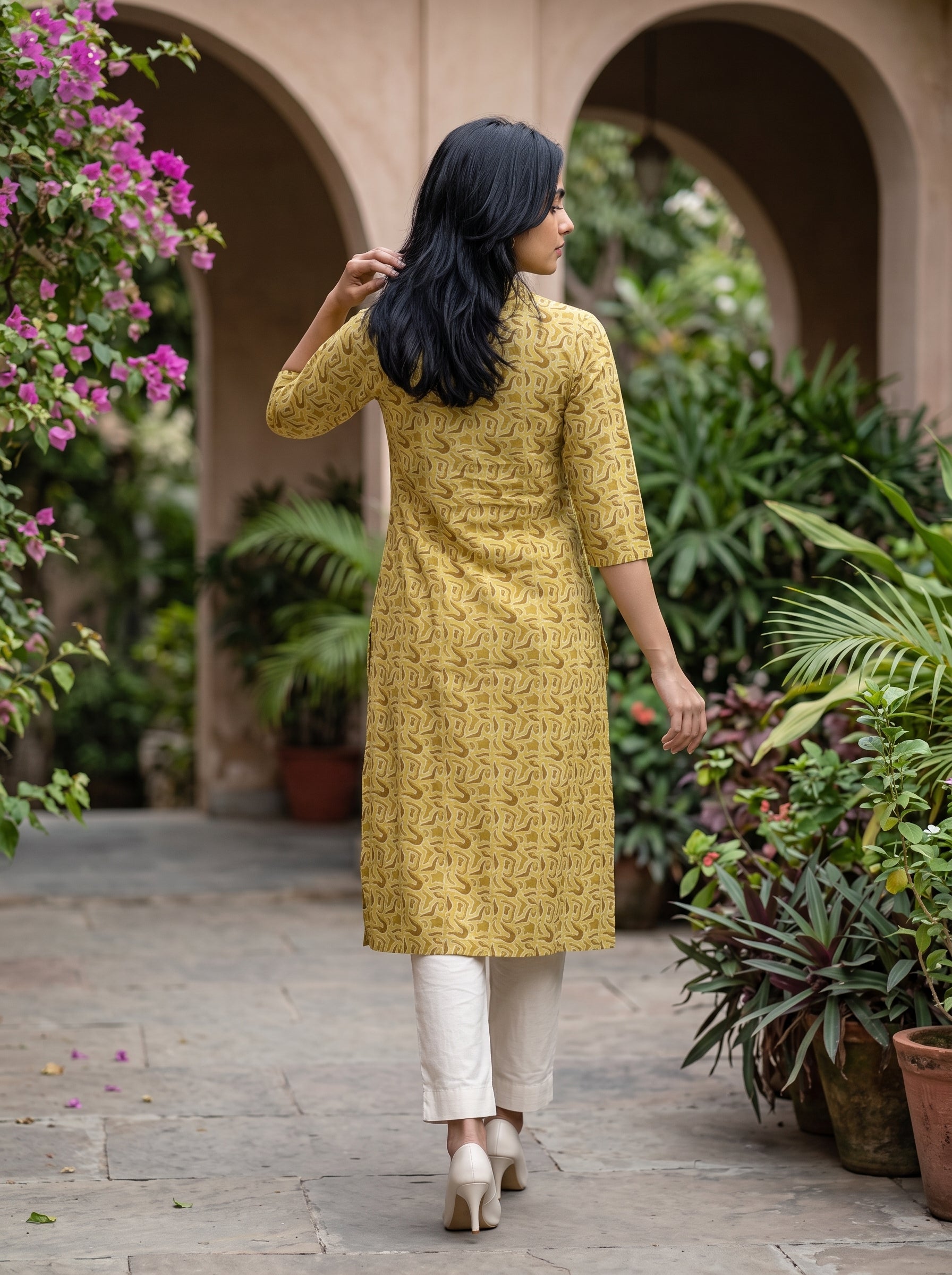 A woman wearing a yellow printed kurta and white pants walks away in a garden courtyard with arches and potted plants.