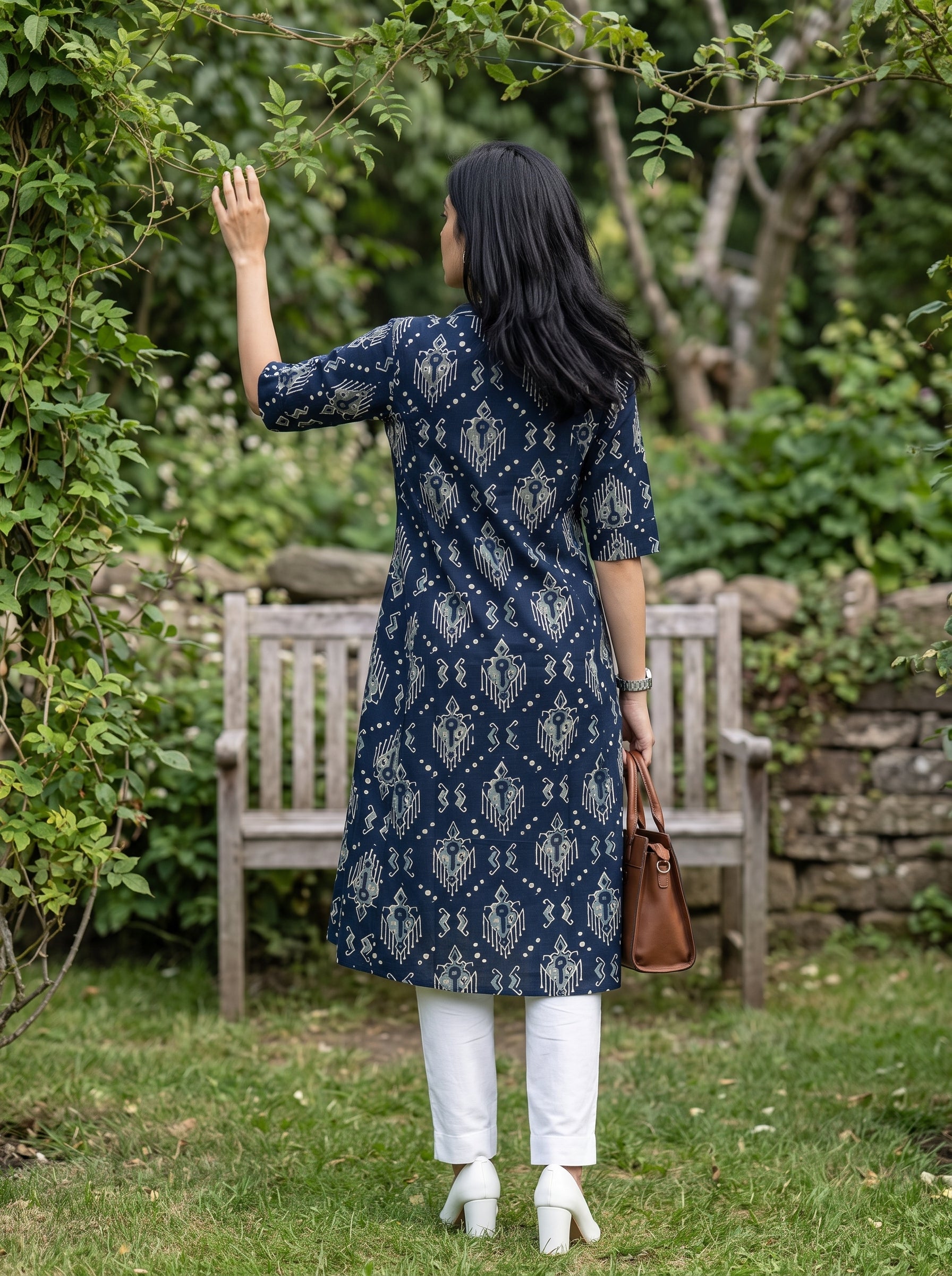 A woman stands in a garden wearing a navy blue and white patterned tunic dress, white pants, and white heels, holding a brown handbag.
