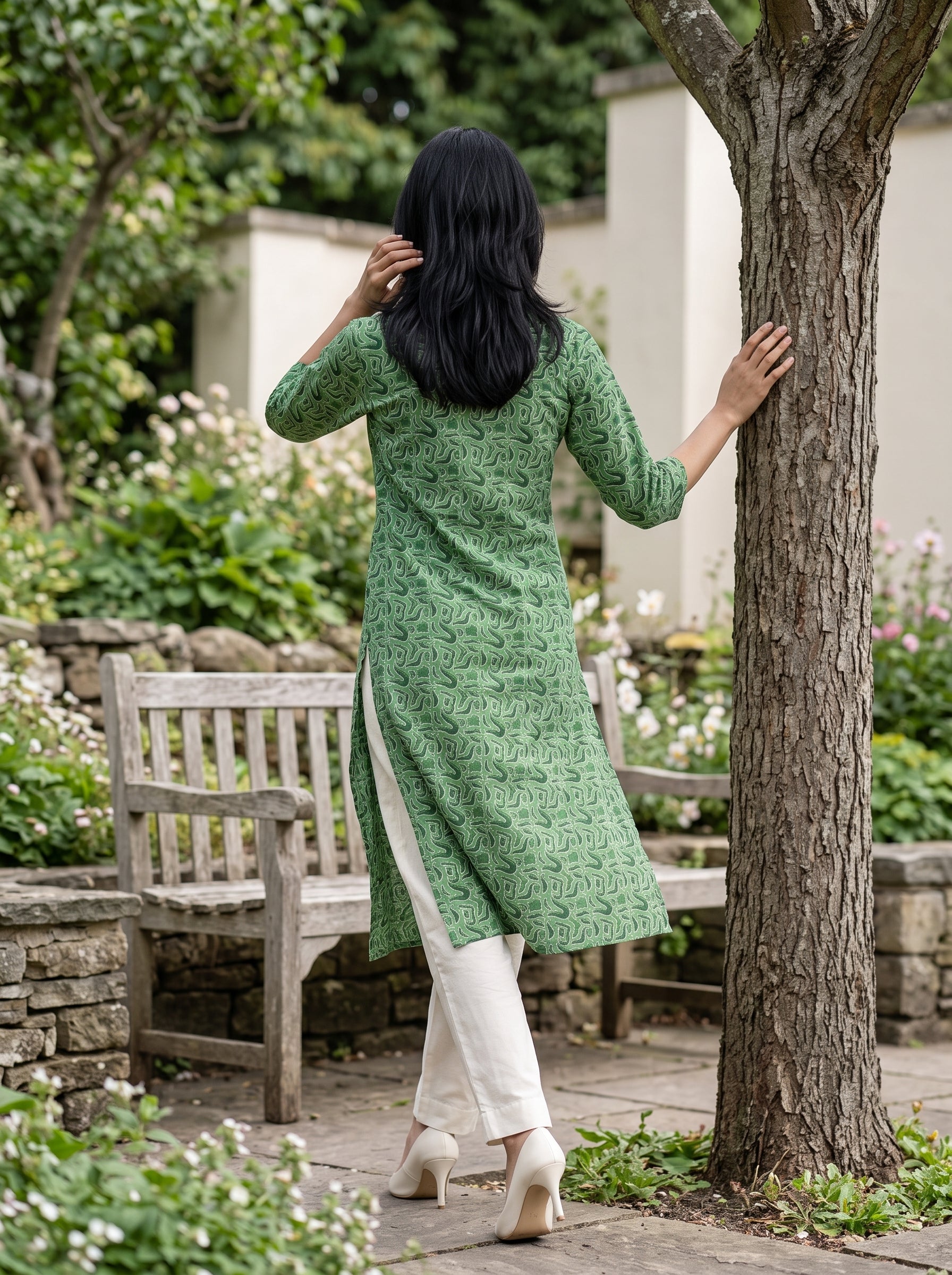 A woman viewed from behind wearing a green patterned kurta and white trousers, standing in a garden next to a tree.