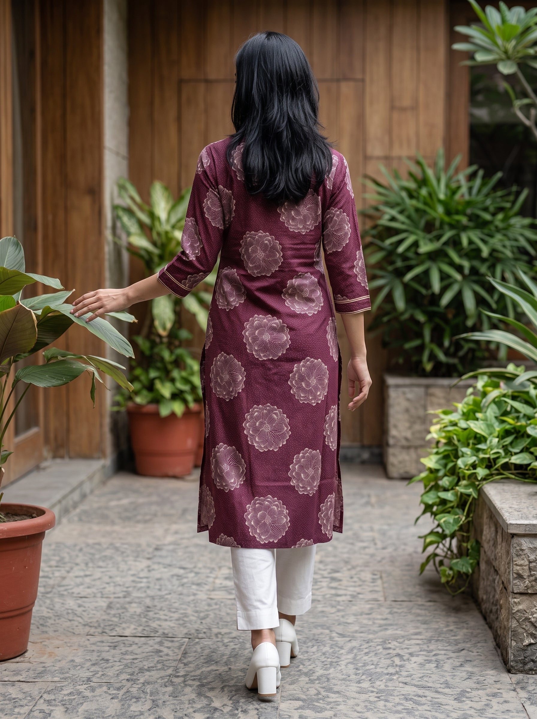 A woman viewed from behind wearing a maroon purple kurta with a beige floral pattern, paired with white pants and white shoes, walking in a garden courtyard.