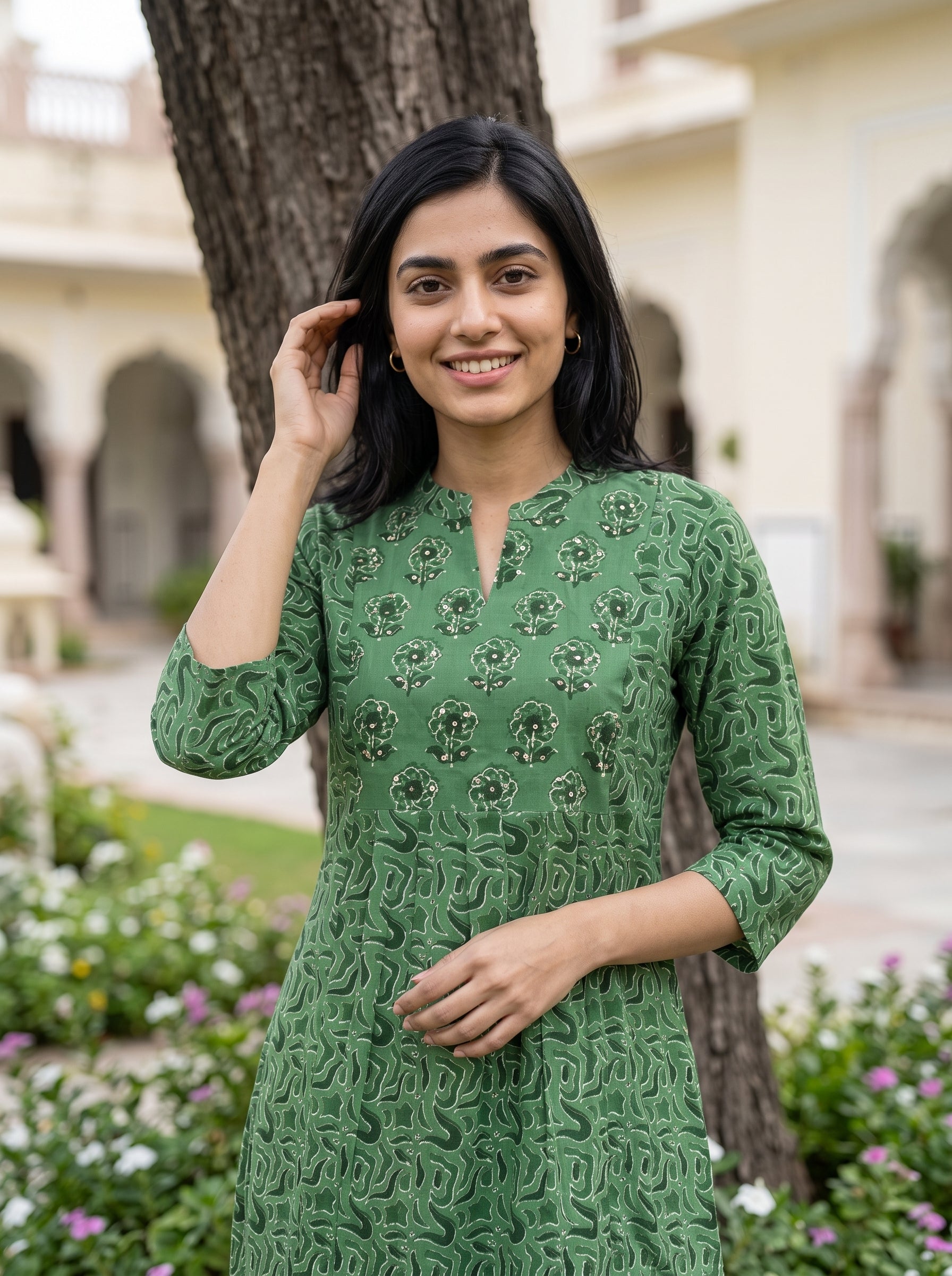 A woman wearing a green embroidered kurta stands outdoors in front of a tree and a building with arches. The kurta has a textured, raised pattern and floral embroidery.
