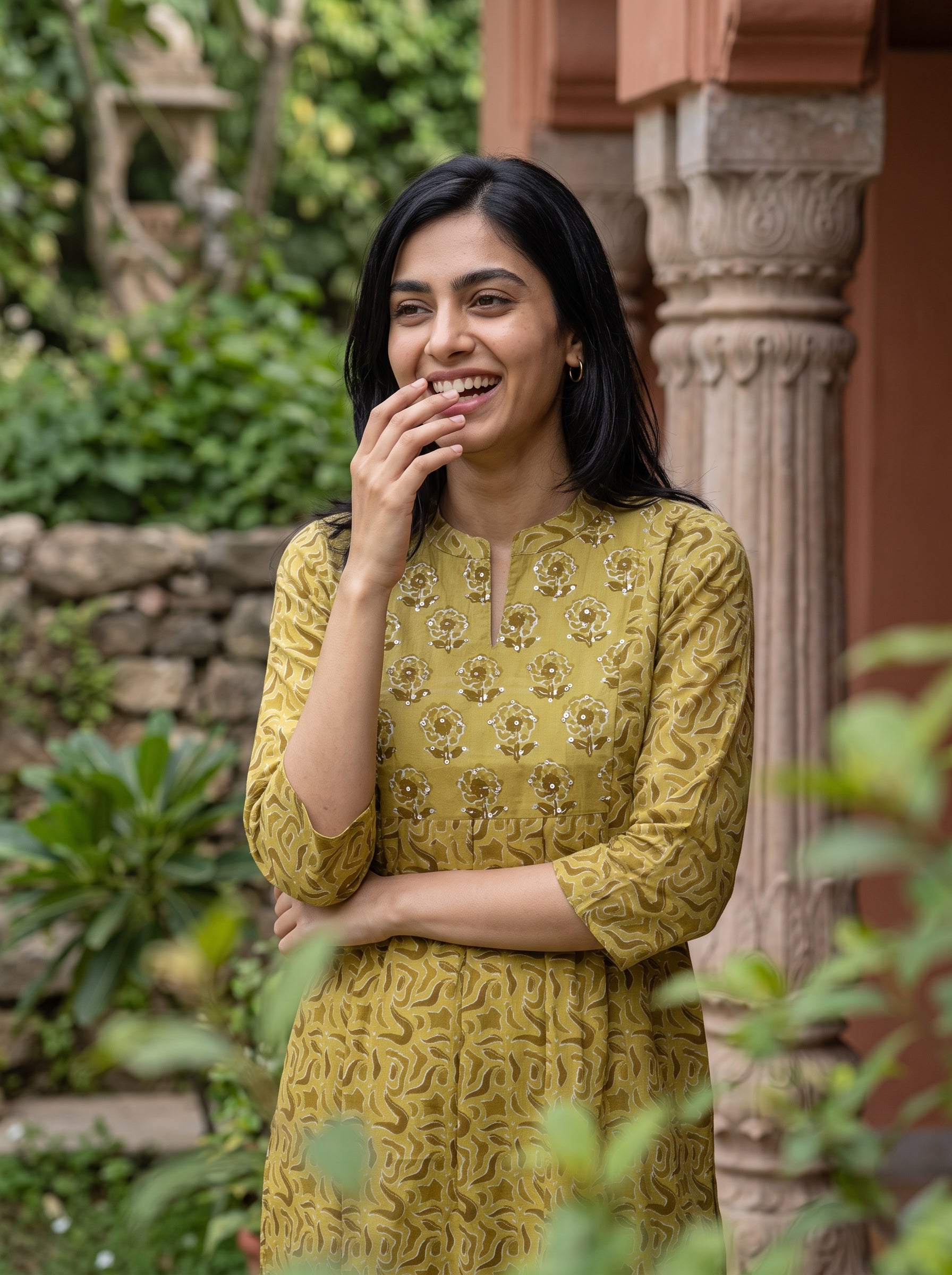 A woman wearing a mustard yellow printed kurta stands outdoors in front of a stone pillar and green foliage, smiling and covering part of her mouth with her hand.