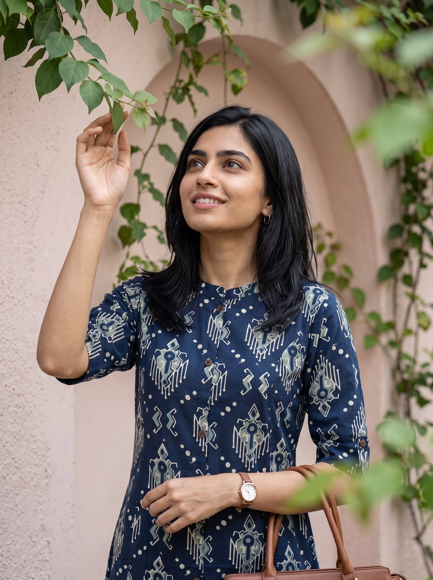 A woman wearing a navy blue short-sleeve kurta with a white geometric block print pattern, standing outdoors against a beige stucco wall with green vines.
