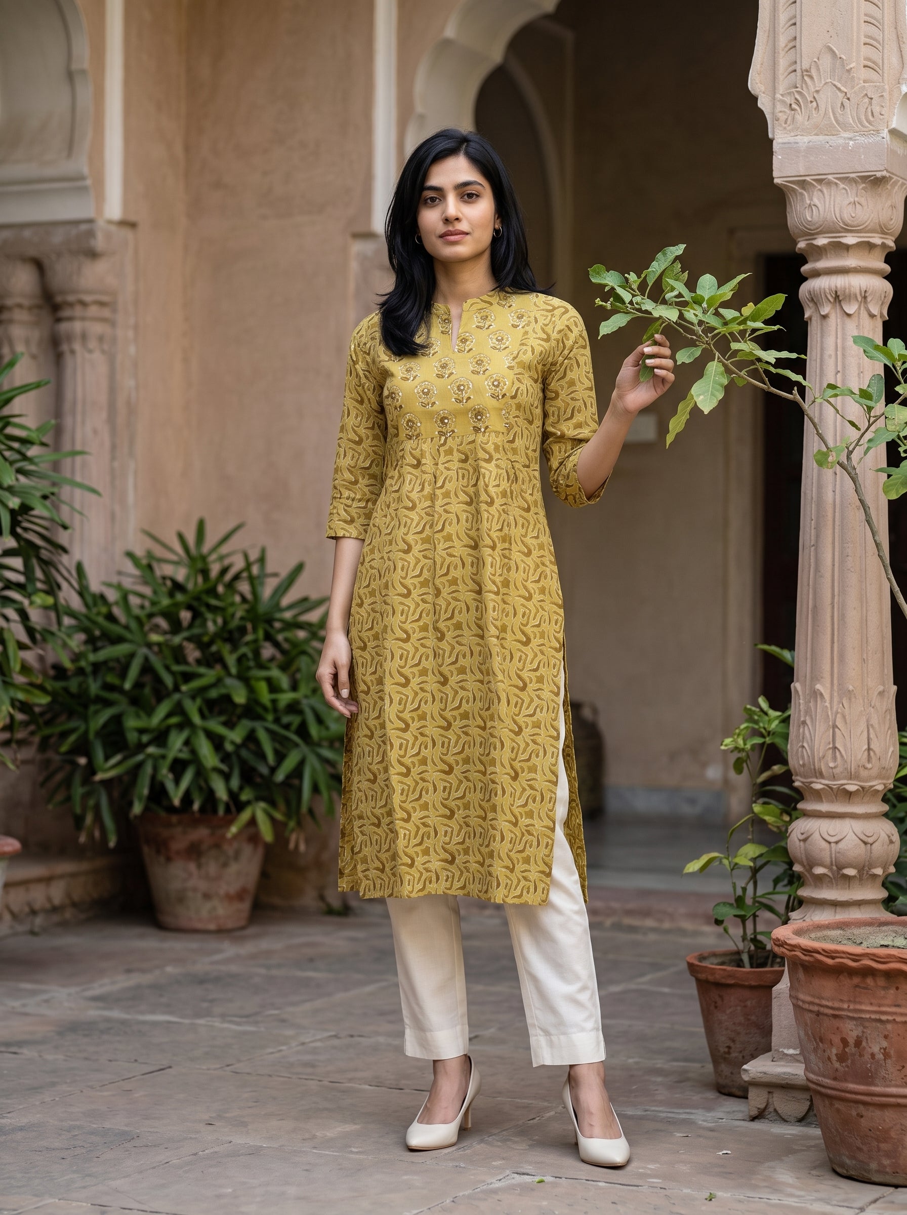 A woman stands in a courtyard wearing a mustard yellow printed kurta with three-quarter sleeves and white pants. The background features beige stone pillars and potted green plants.