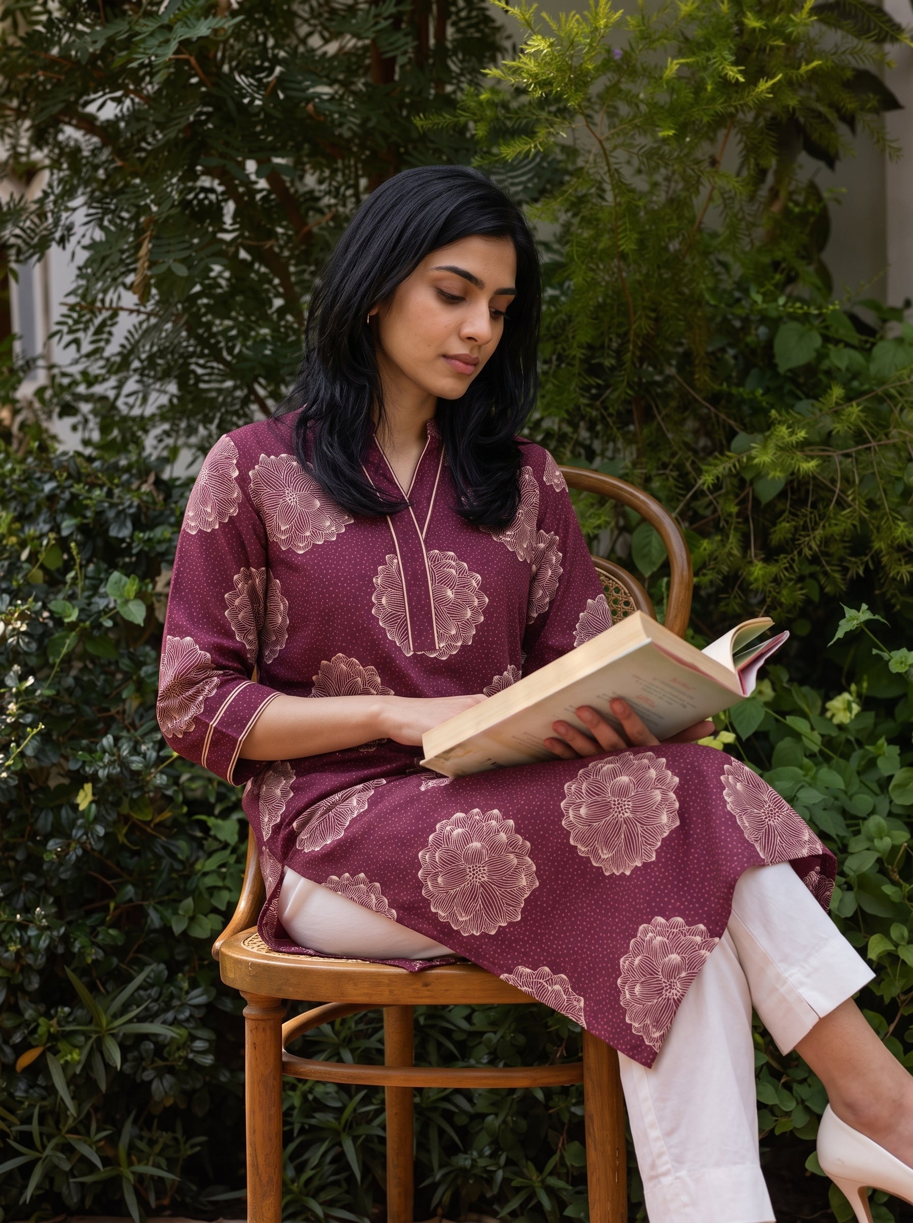A woman sits on a wooden chair outdoors, reading a book. She is wearing a deep purple kurta with a light pink floral pattern and white trousers, set against a background of green foliage.