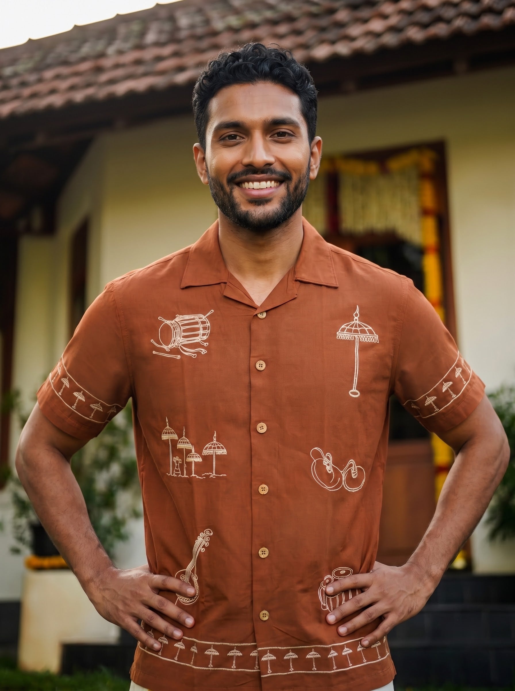 A man wearing a rust-orange short-sleeve button-up shirt with white tropical and cultural motifs, standing outdoors in front of a building with a tiled roof.