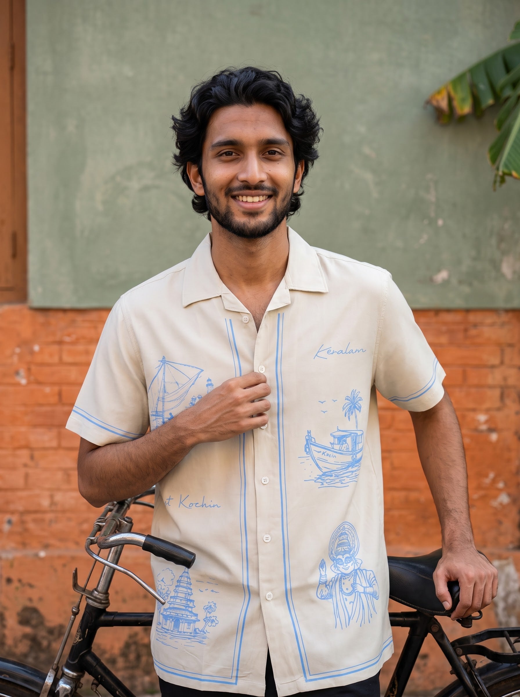 A man wearing a cream-colored short-sleeve button-up shirt with light blue illustrations and text, standing next to a bicycle in front of a rustic brick and stucco wall.