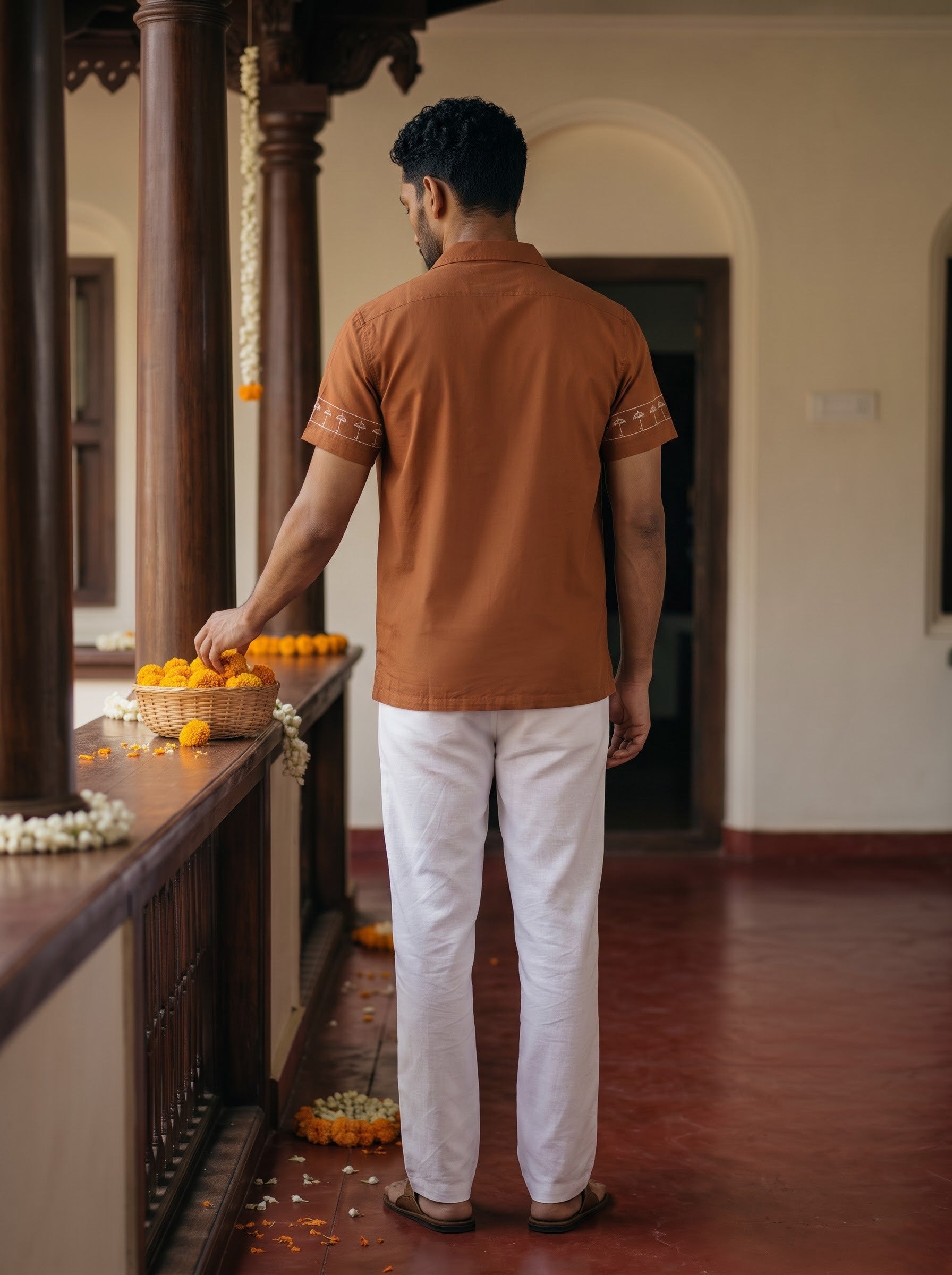 A man viewed from behind wearing a rust-orange short-sleeve kurta with white trousers, standing on a traditional porch with wooden pillars and a basket of oranges on the railing.