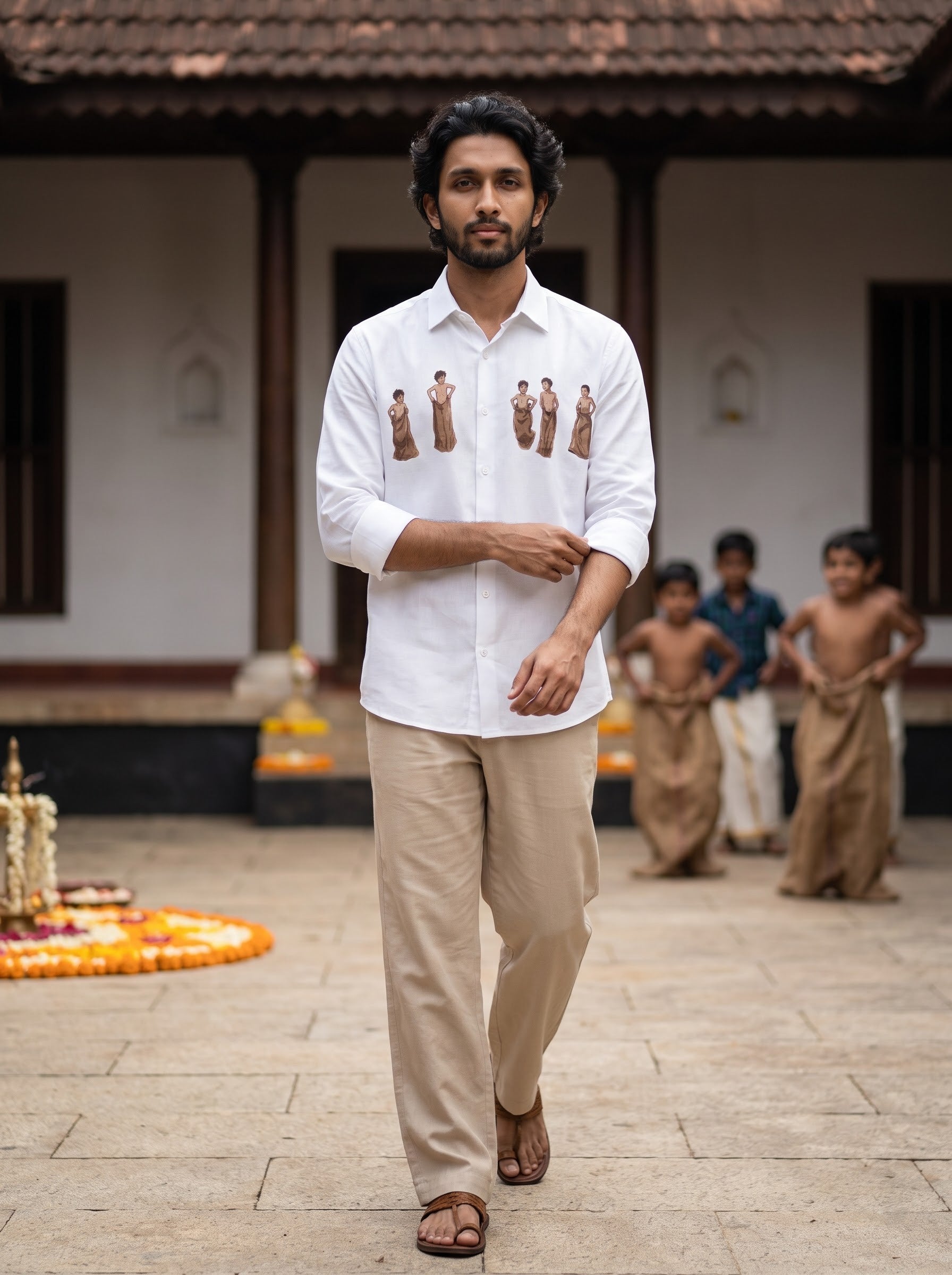 A man walking outdoors in front of a traditional building, wearing a white long-sleeve button-up shirt with small brown figurine prints on the chest, paired with beige trousers and brown sandals. Children are visible in the background.