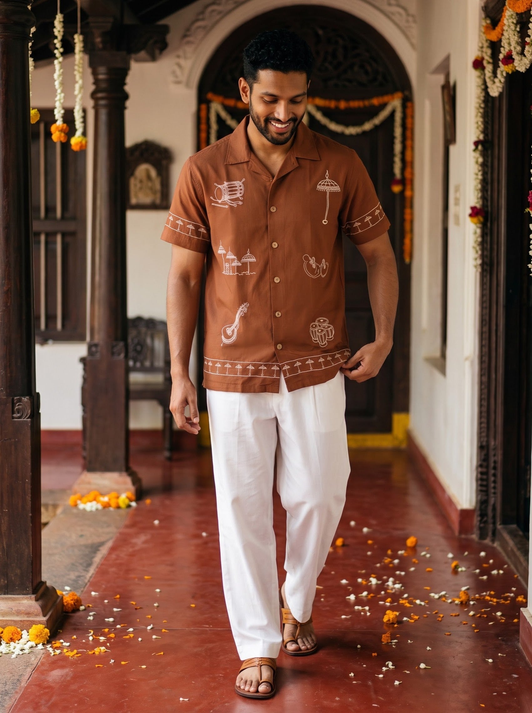 A man walking down a decorated corridor wearing a brown short-sleeve button-up shirt with white traditional Indian prints, paired with white trousers and brown sandals.