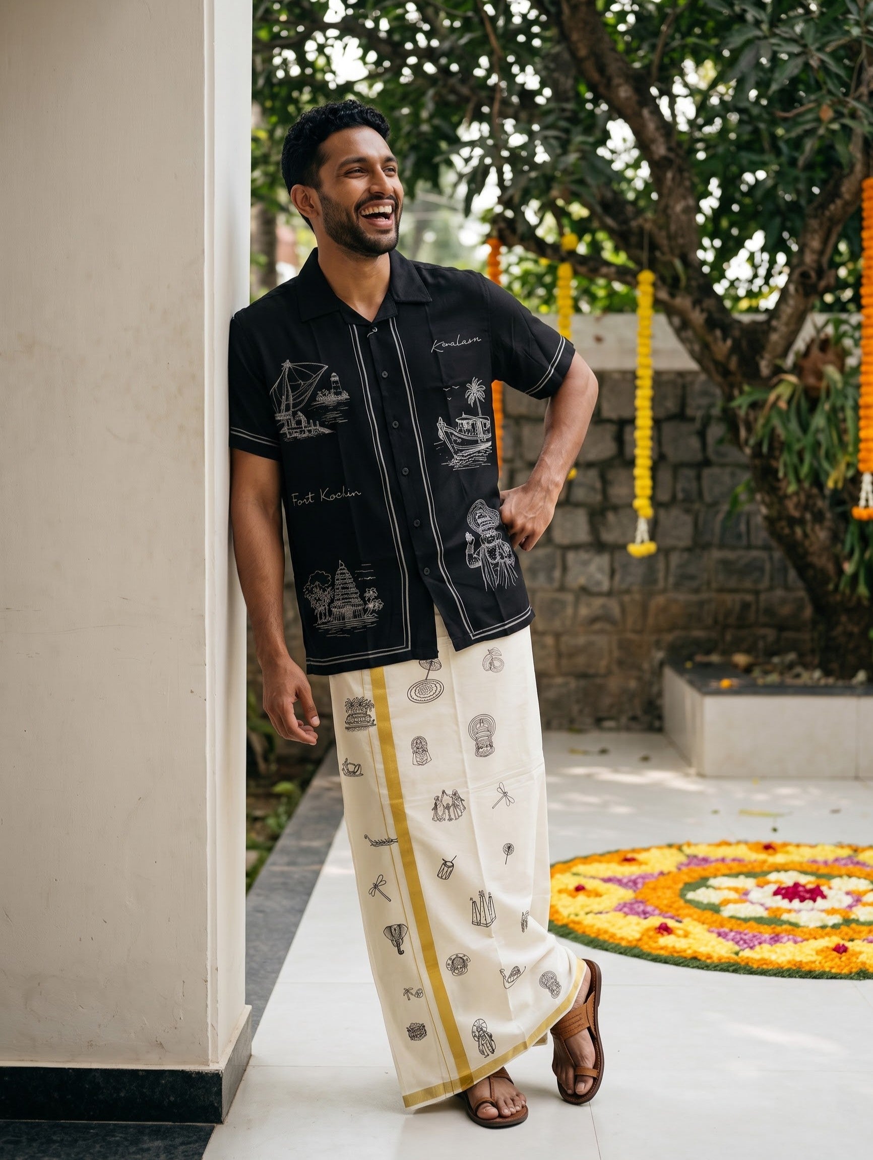 A man wearing a black short-sleeve button-up shirt with white illustrations and a cream-colored traditional wrap skirt with yellow and black prints, standing in an outdoor courtyard.