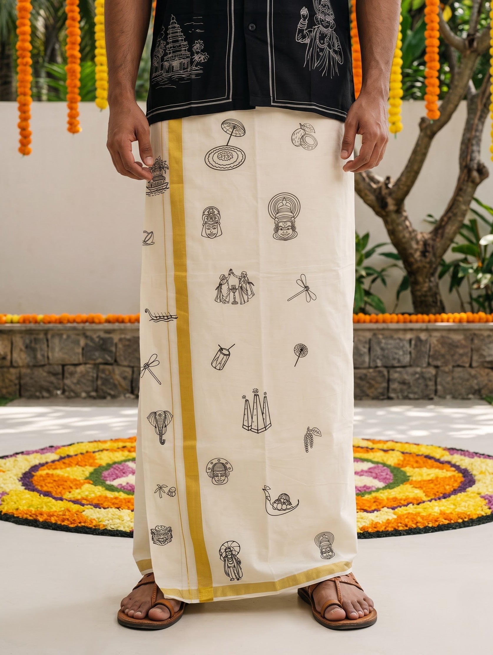 A man wearing a traditional cream-colored cotton lungi skirt with black printed motifs and a yellow stripe down the side, paired with a black shirt and brown sandals, standing outdoors on a decorated floor.