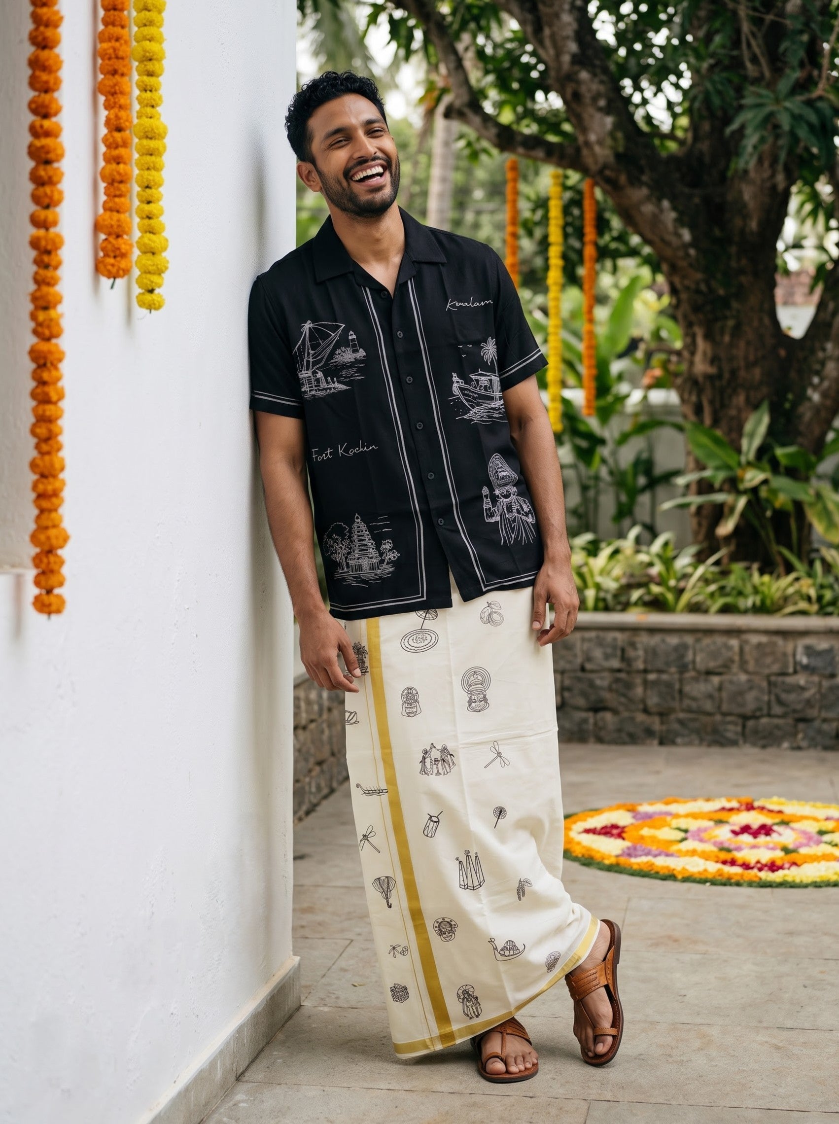 A man wearing a black short-sleeve button-up shirt with white illustrations and a cream-colored traditional wrap skirt with yellow stripes and printed motifs, standing outdoors against a white wall.
