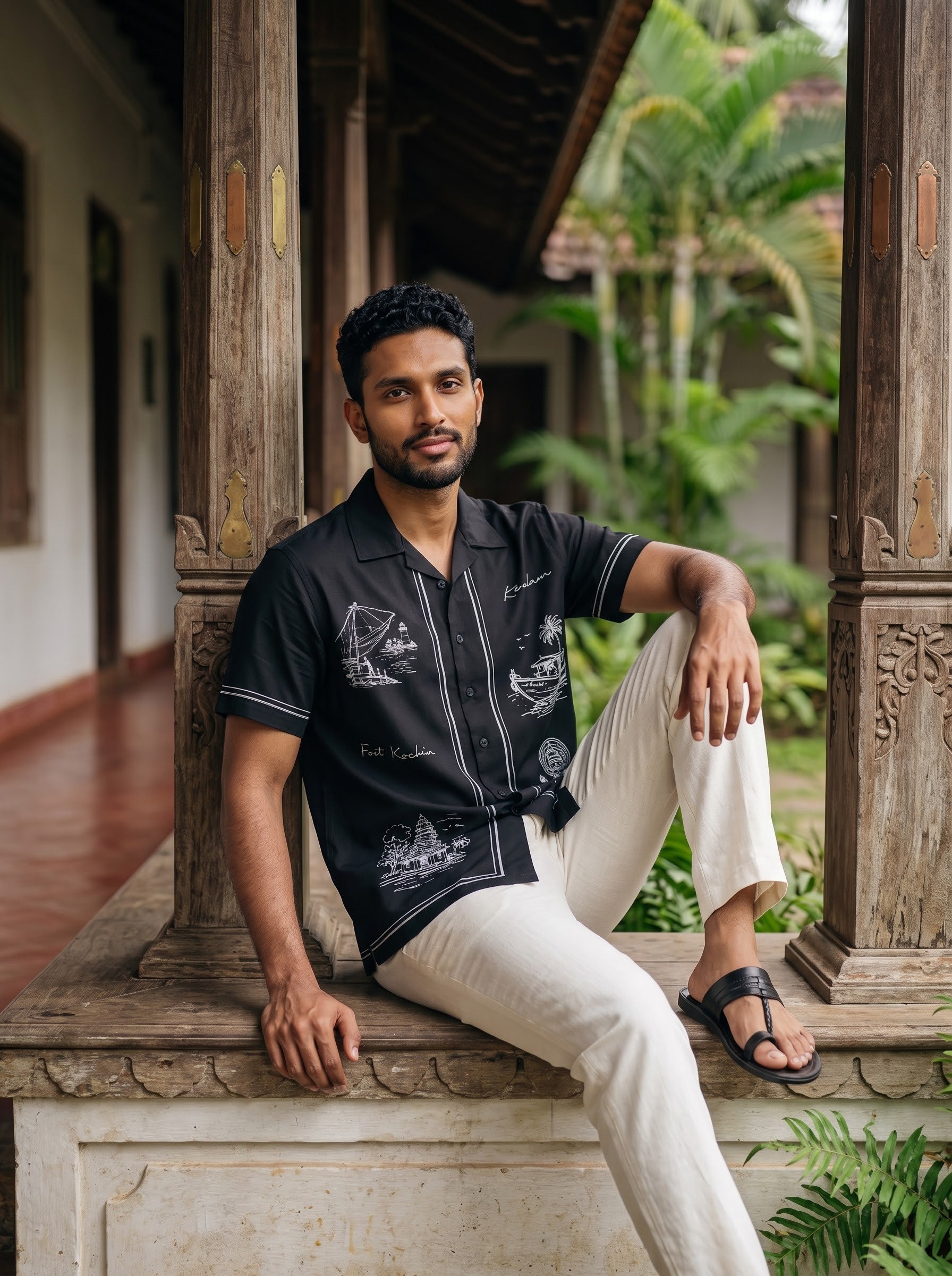 A man sitting on a stone ledge in a traditional courtyard, wearing a black short-sleeve button-up shirt with white boat illustrations and cream-colored pants.