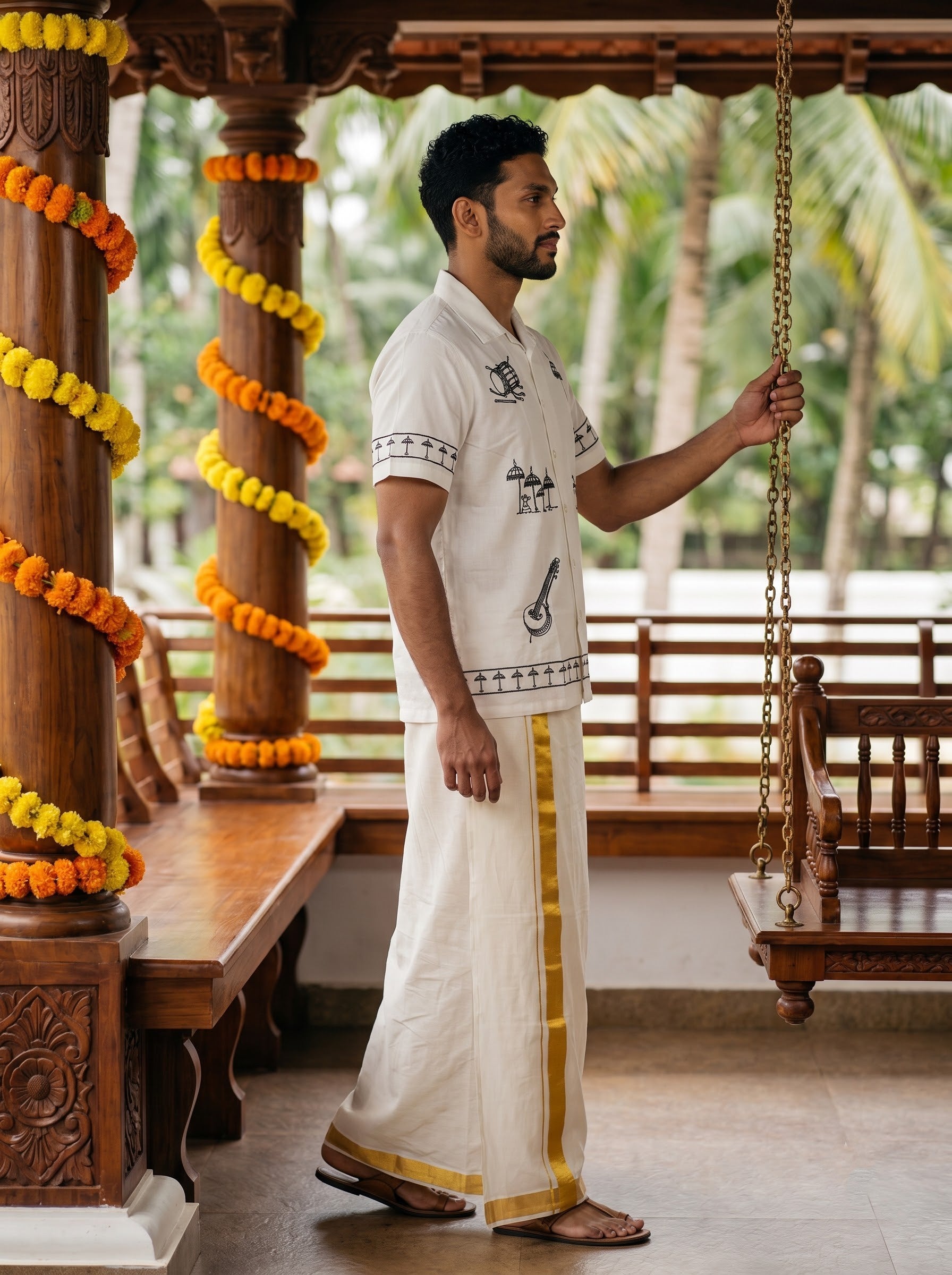 A man wearing a traditional white Tamil kurta and dhoti with gold borders stands in a wooden porch, holding a hanging swing. The porch is decorated with orange and yellow marigold garlands.