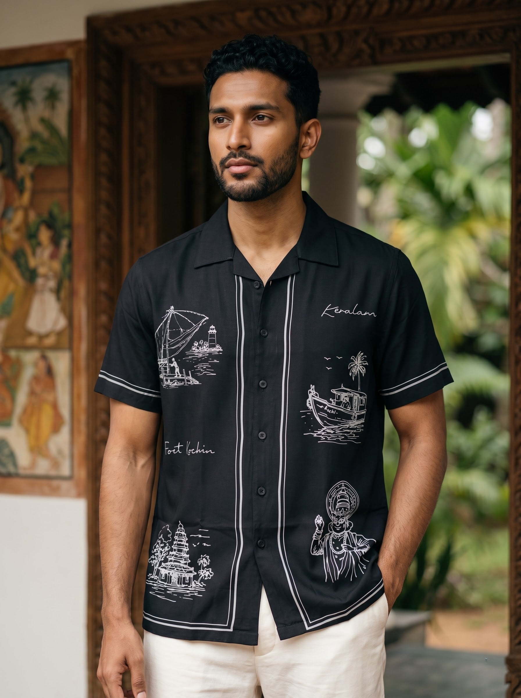 A man wearing a black short-sleeve button-up shirt with white tropical boat and cultural illustrations, paired with off-white trousers, standing in front of a wooden doorway with green foliage in the background.