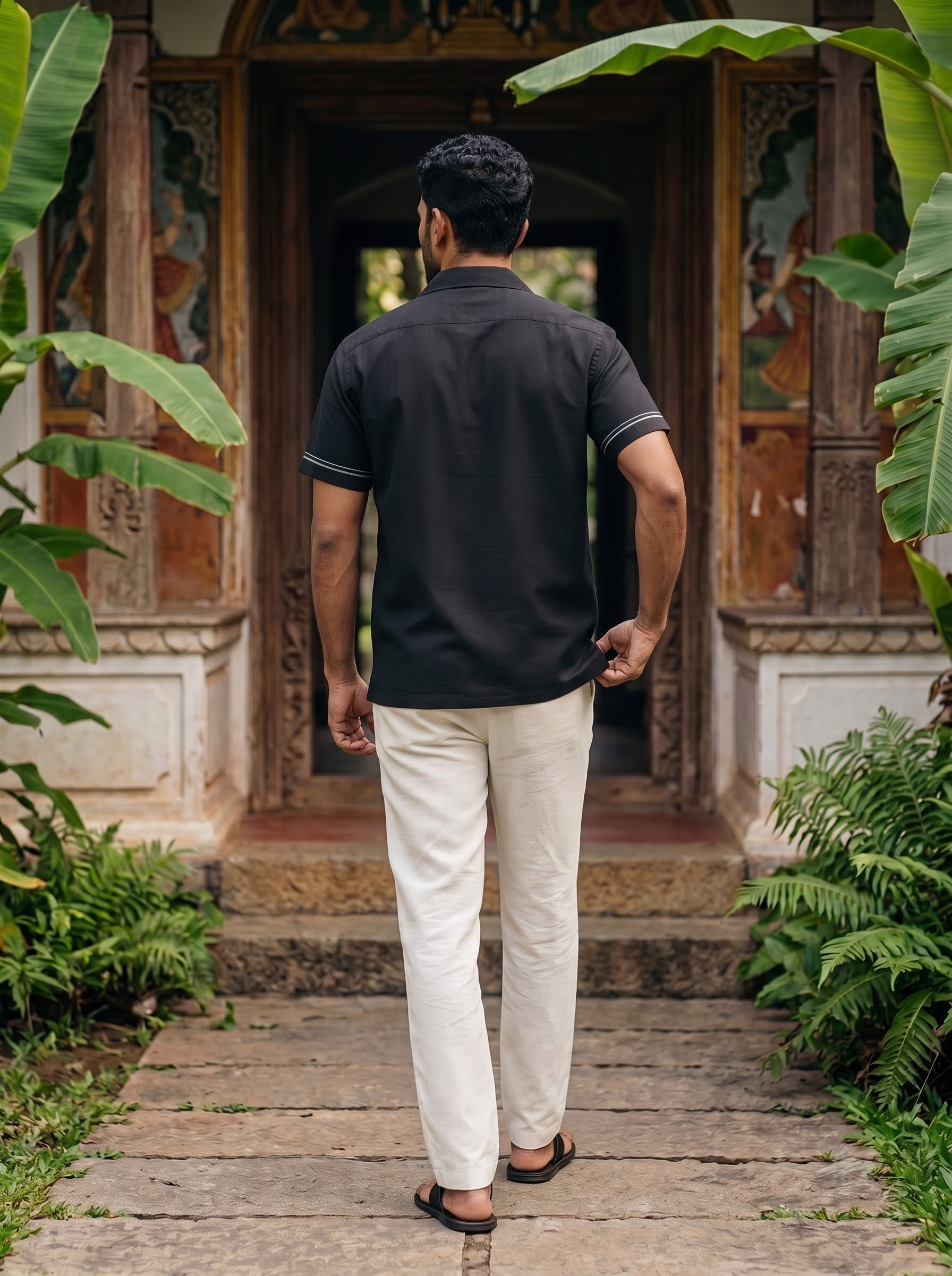 A man viewed from behind wearing a black short-sleeve polo shirt with white stripe accents on the collar and sleeve cuffs, paired with off-white trousers, standing on a stone path in front of a traditional wooden doorway surrounded by lush green foliage.
