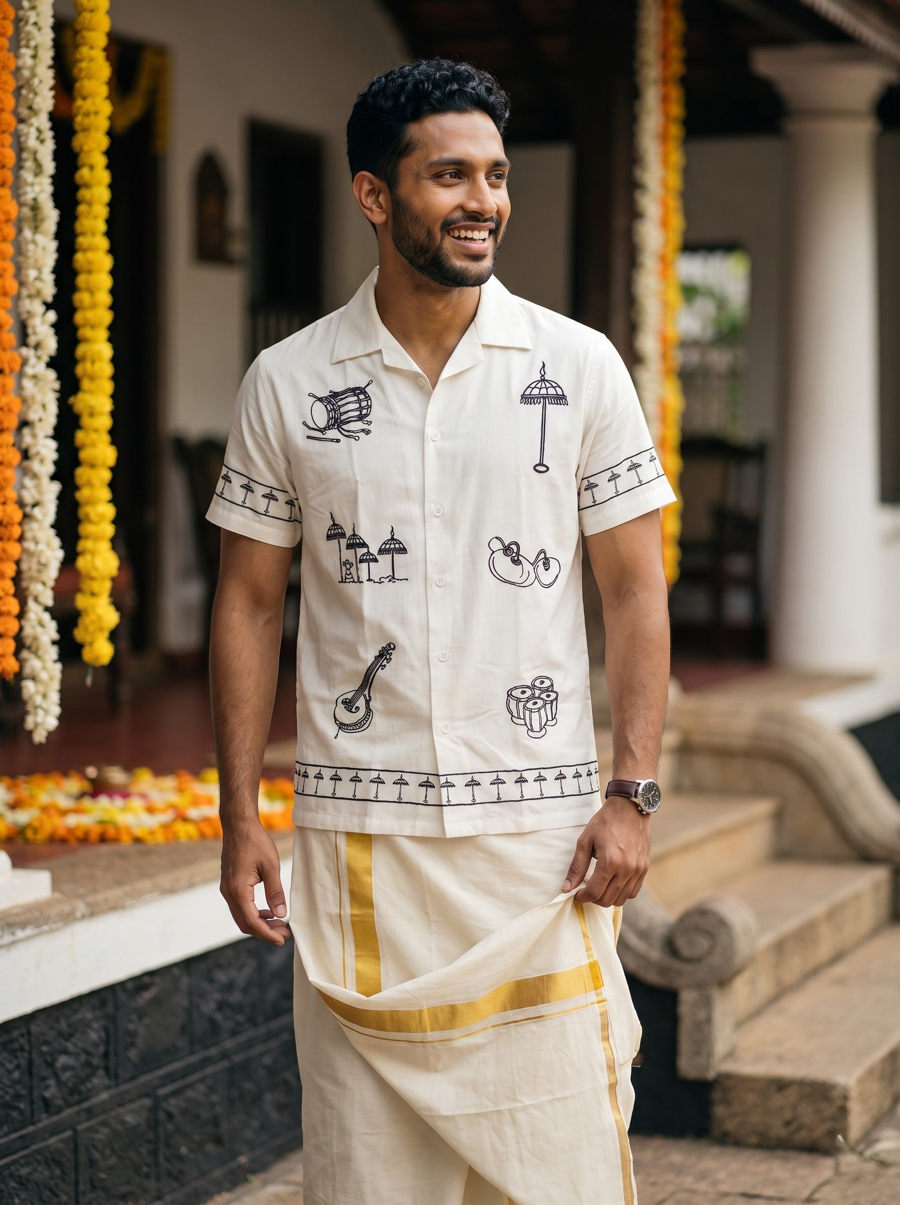 A man wearing a traditional Indian outfit consisting of a cream-colored short-sleeve kurta with black embroidered motifs and a matching drape, standing in front of a building with garlands.