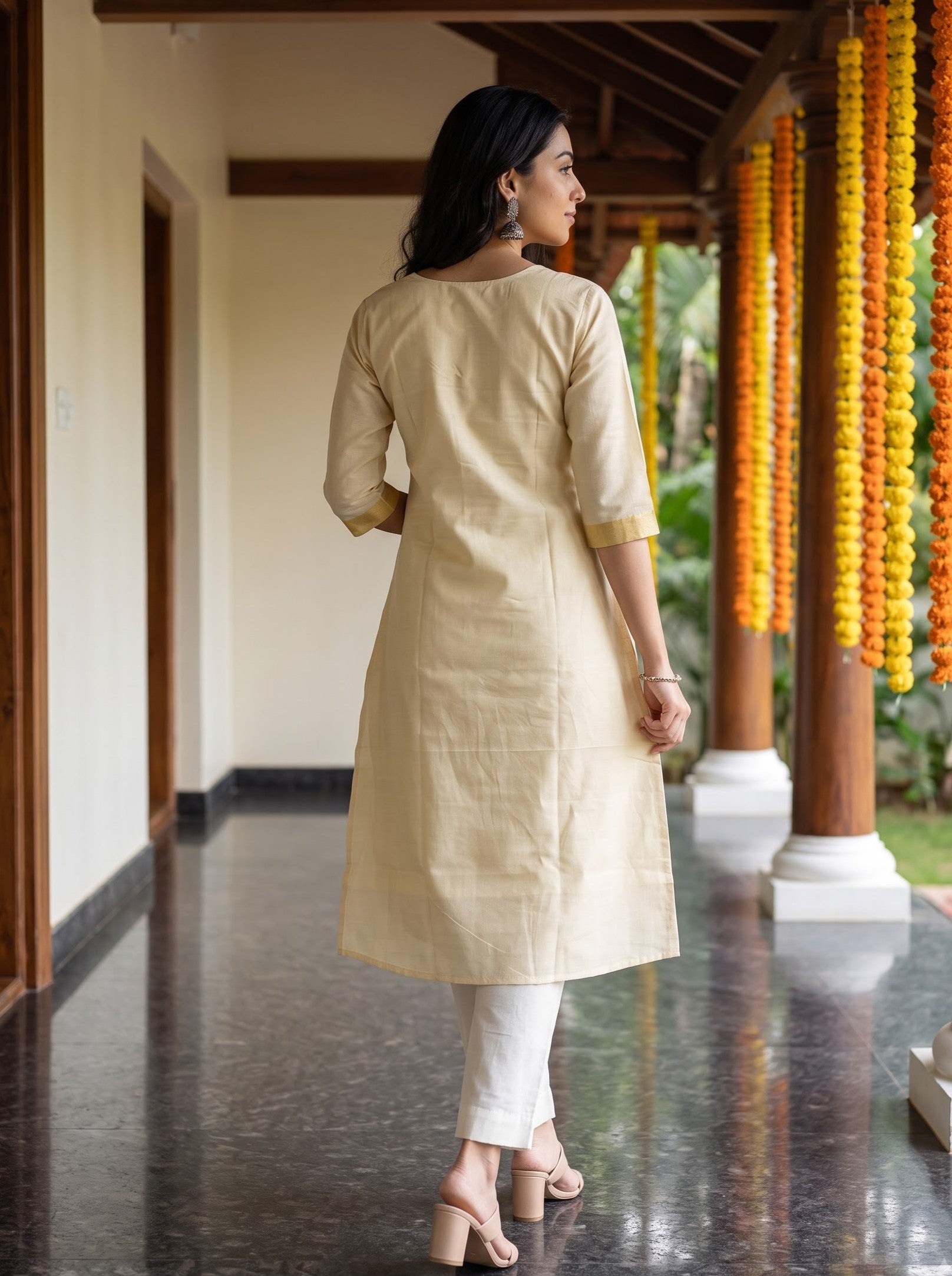 A woman wearing a light beige linen kurta with white pants and beige heels, standing on a covered walkway with yellow marigold garlands in the background.