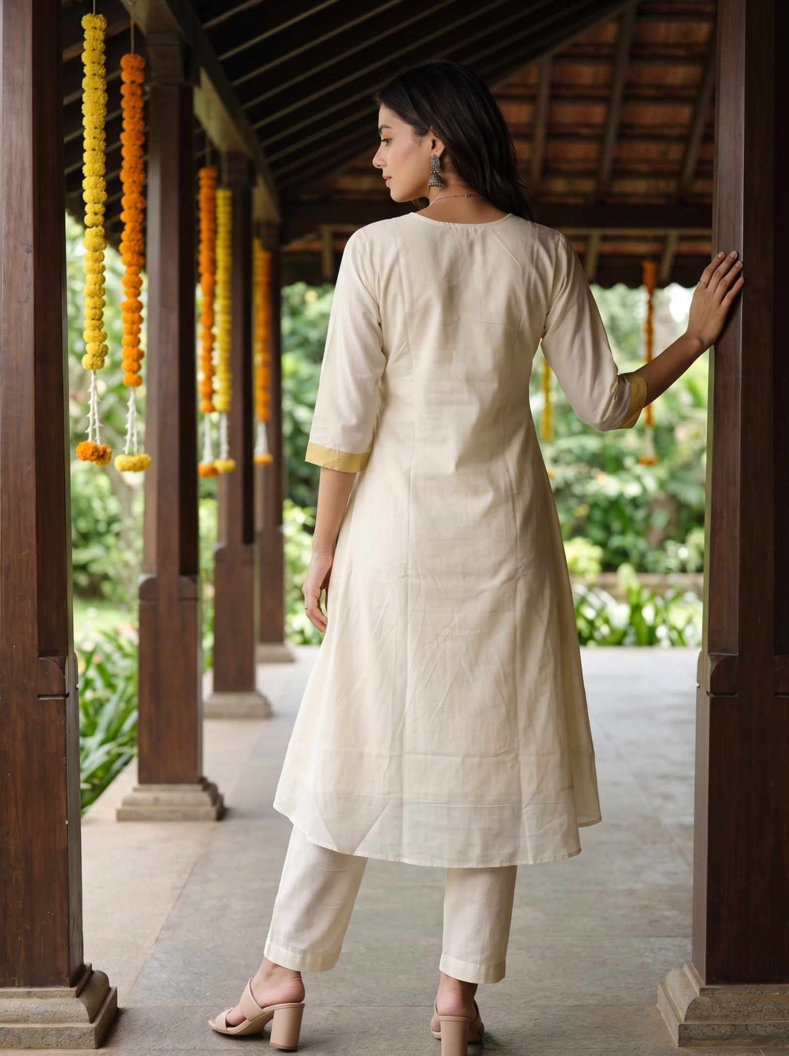 A woman wearing a light beige kurta and matching trousers stands in a traditional wooden corridor, viewed from behind. The kurta has three-quarter sleeves with a subtle yellow-green contrast trim at the cuffs.