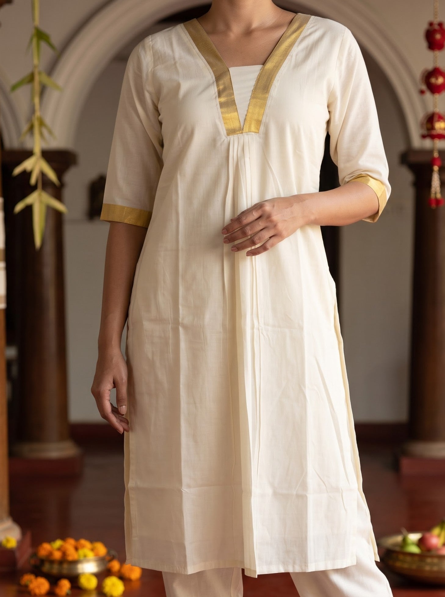 A woman wearing a cream-colored cotton kurta with gold trim on the V-neckline and sleeve cuffs, paired with matching cream trousers, set against an indoor background with arches and decorative elements.